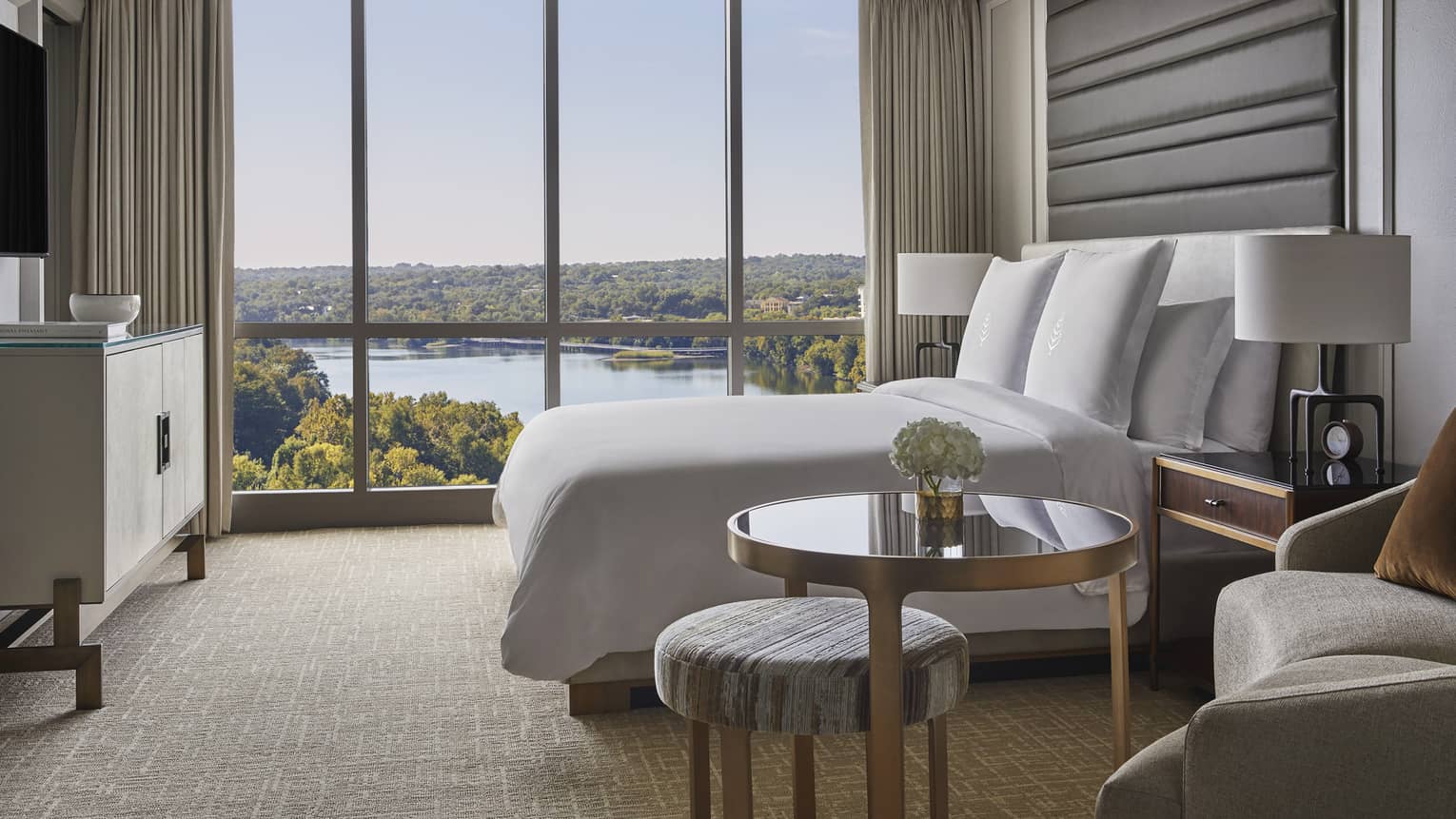 Side view of small brass and glass table, hotel bed by floor to ceiling window and lake views