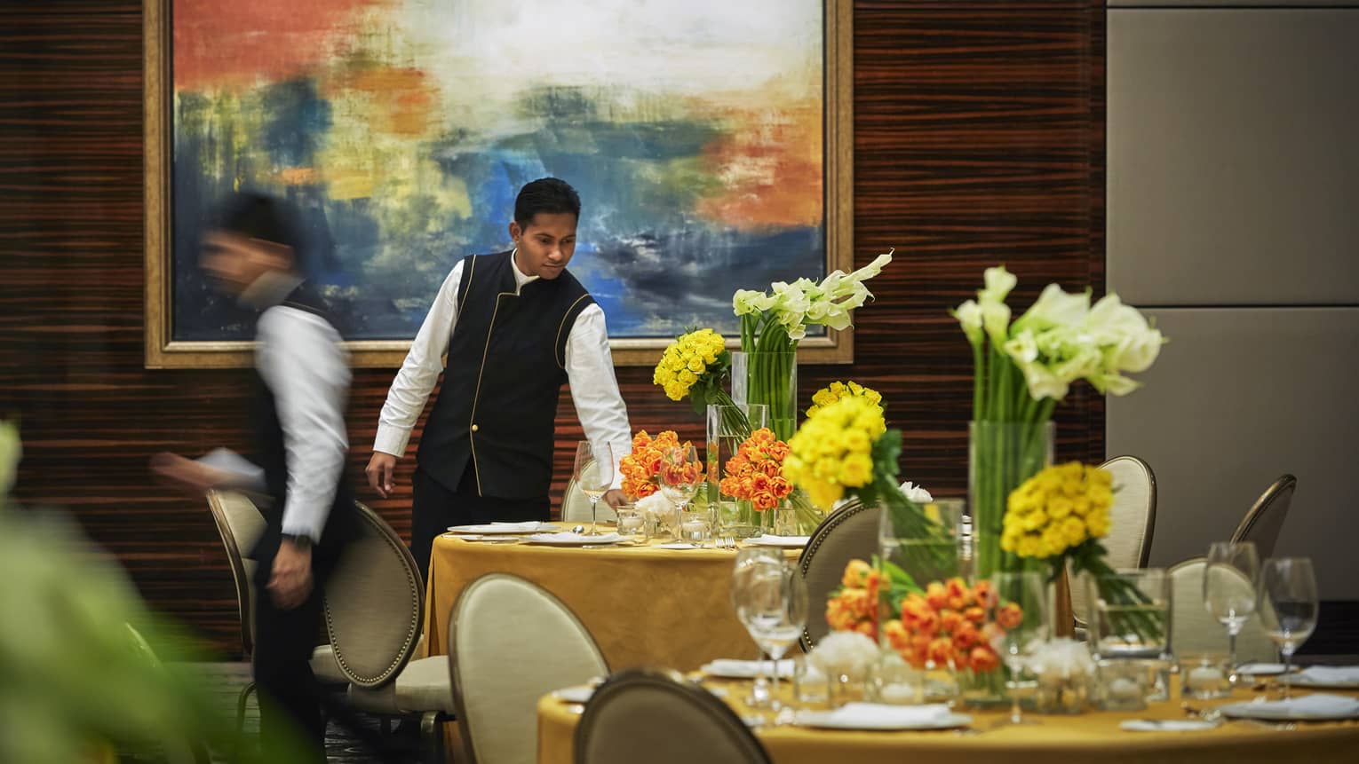 Server in black-and-white uniform sets round formal dining table with yellow linen, vases of fresh orange, yellow flowers