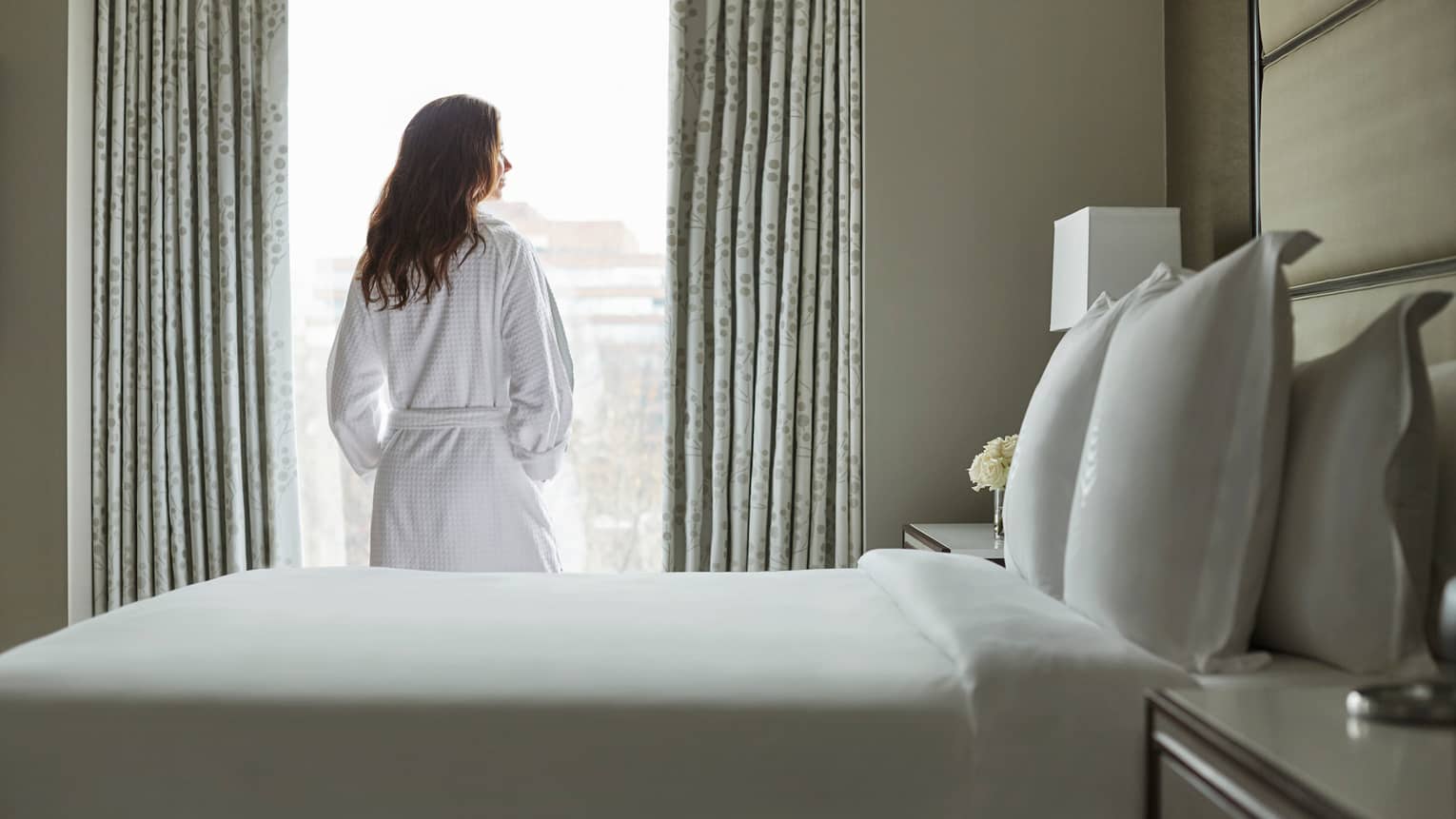 Back of woman in white bathrobe stands at sunny window in front of bed