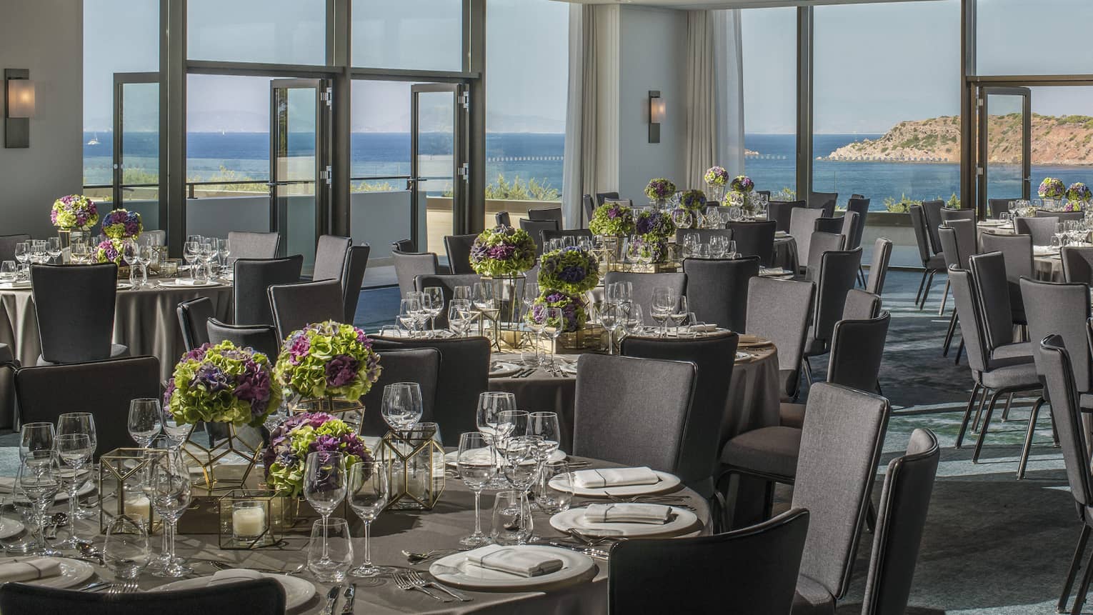Light filled Nafsika Ballroom with round tables, grey chairs, flowers, wine glasses overlooking ocean 