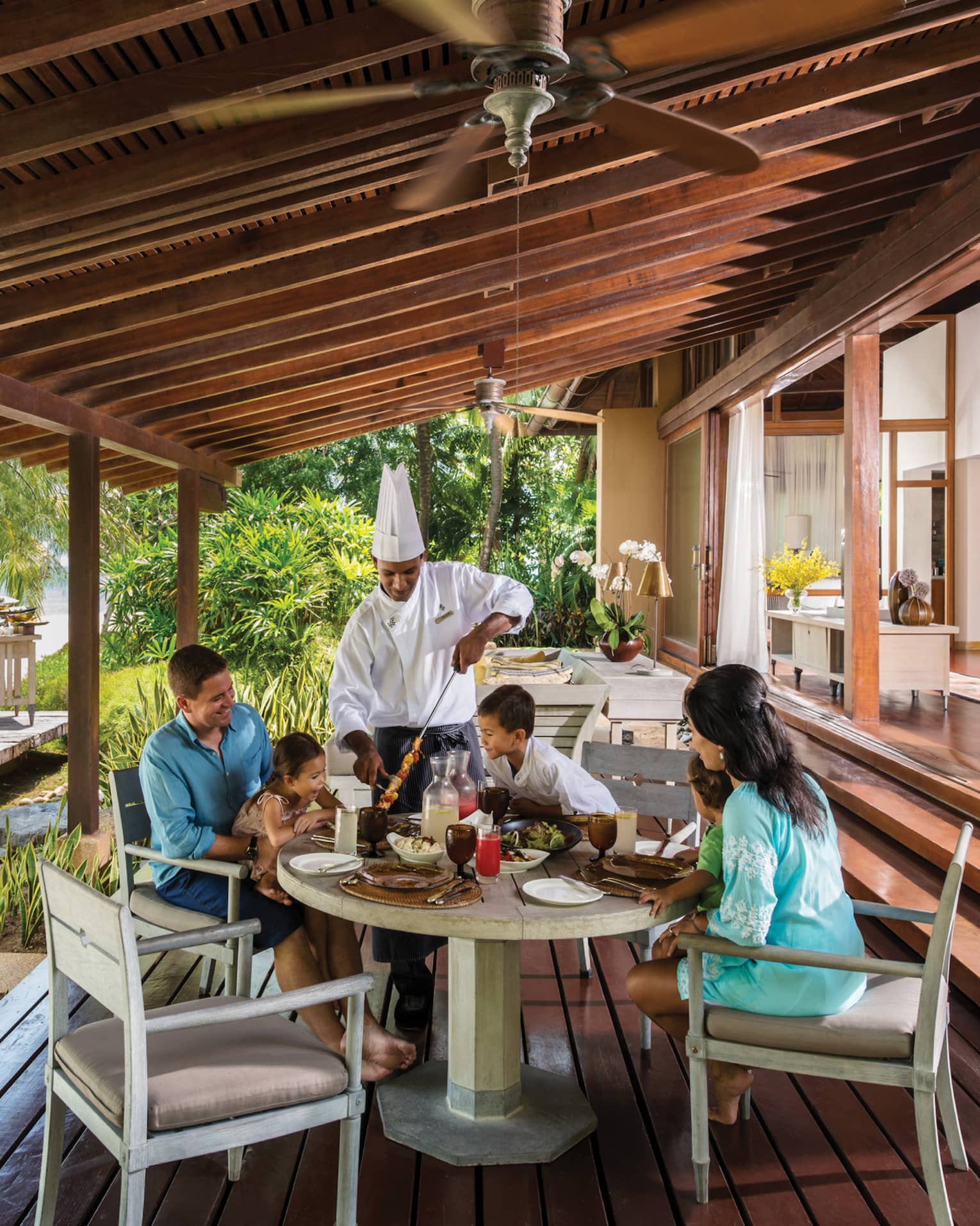 Chef in uniform presents meat skewer to family around table on Beach Villa patio