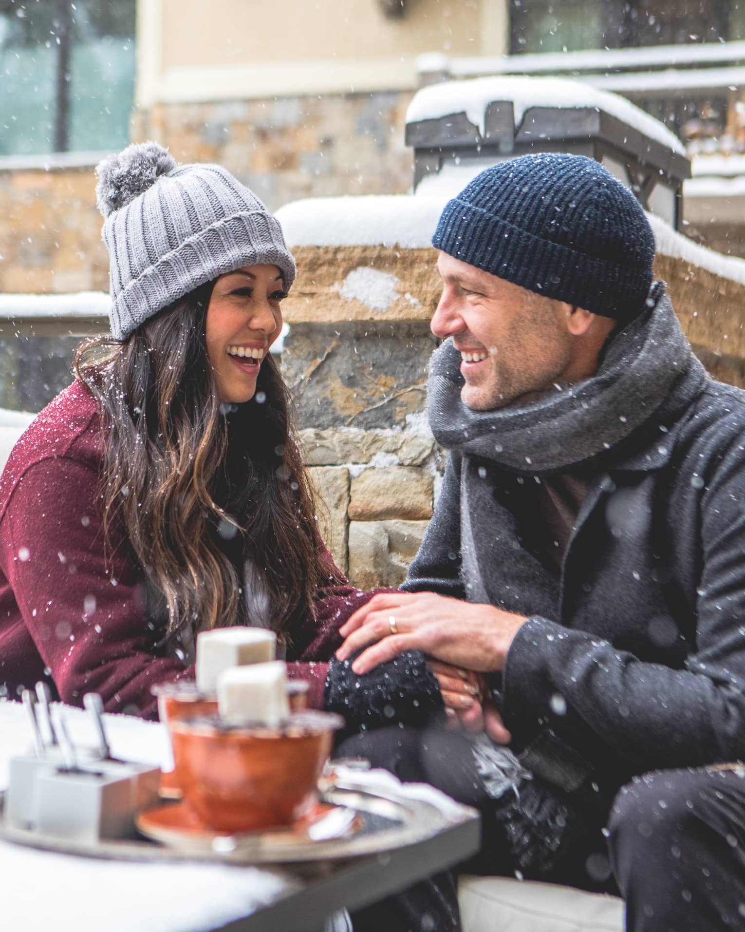 Smiling couple wearing winter clothes sit by hot chocolate tray, modern bonfire fireplace