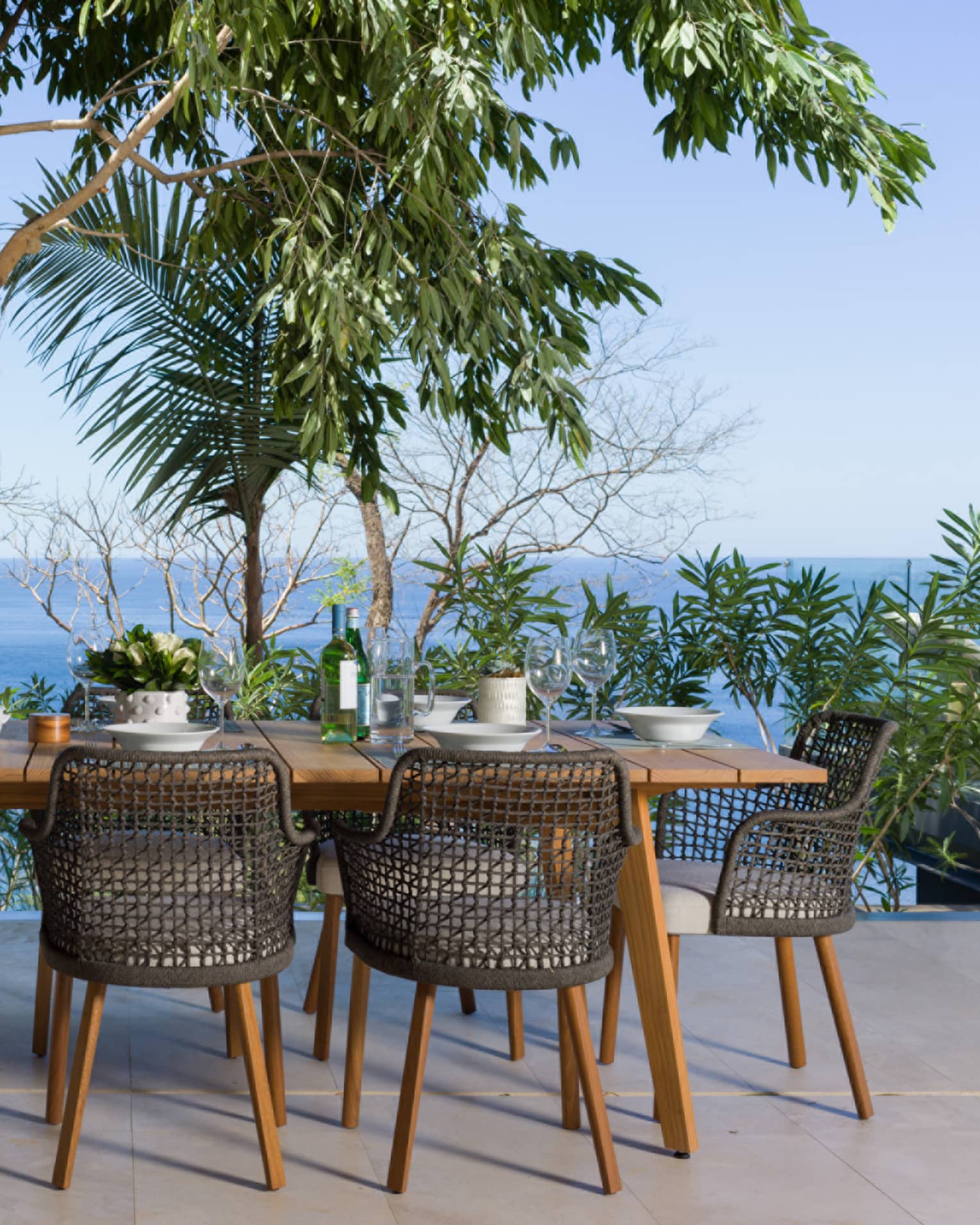 Outdoor shaded dining table set for eight amid lush tropical foliage on a terrace overlooking treetops, vast ocean beyond.