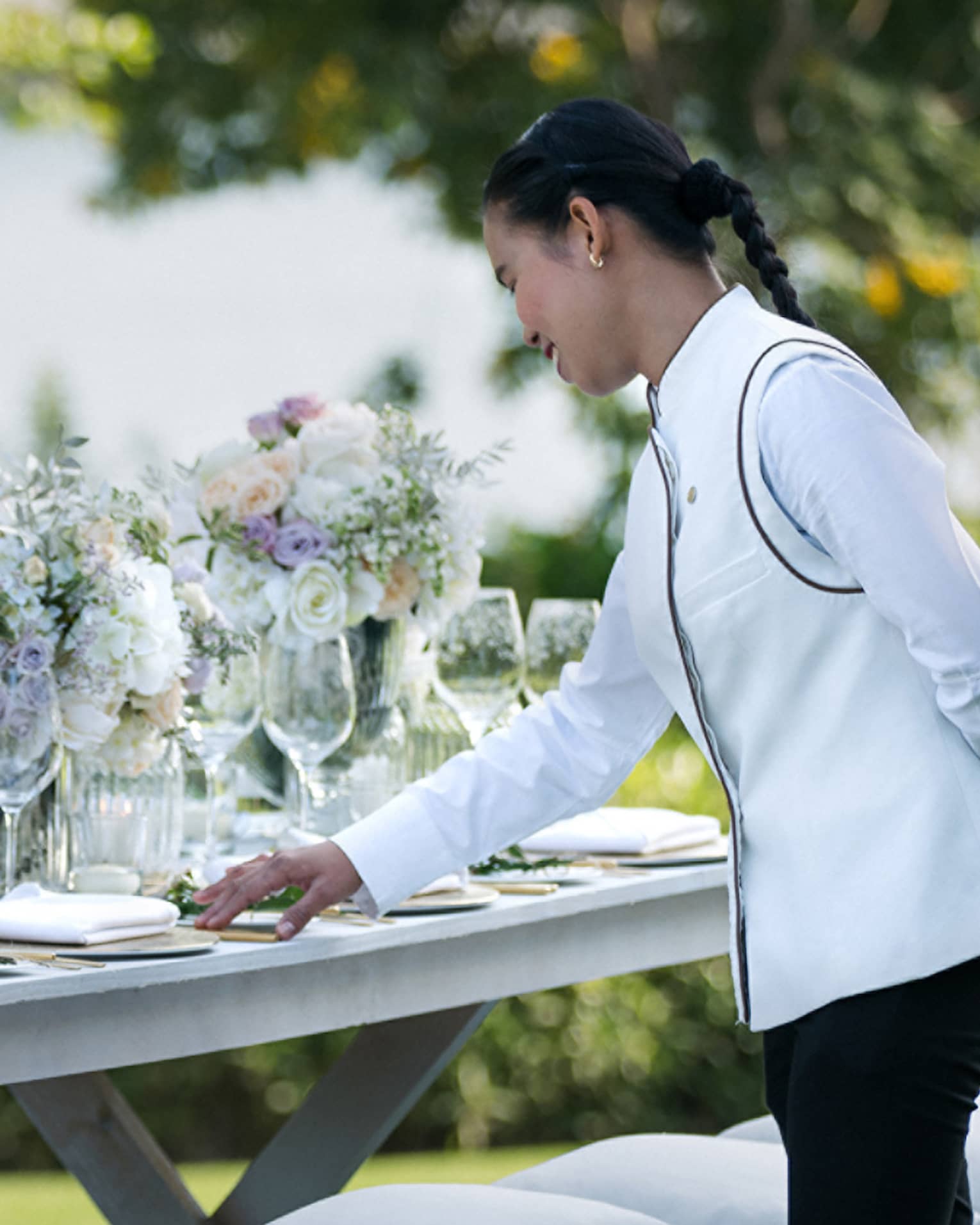 A four seasons staff does the final touches on an outdoor, long banquet table decorated with large white floral arrangements 