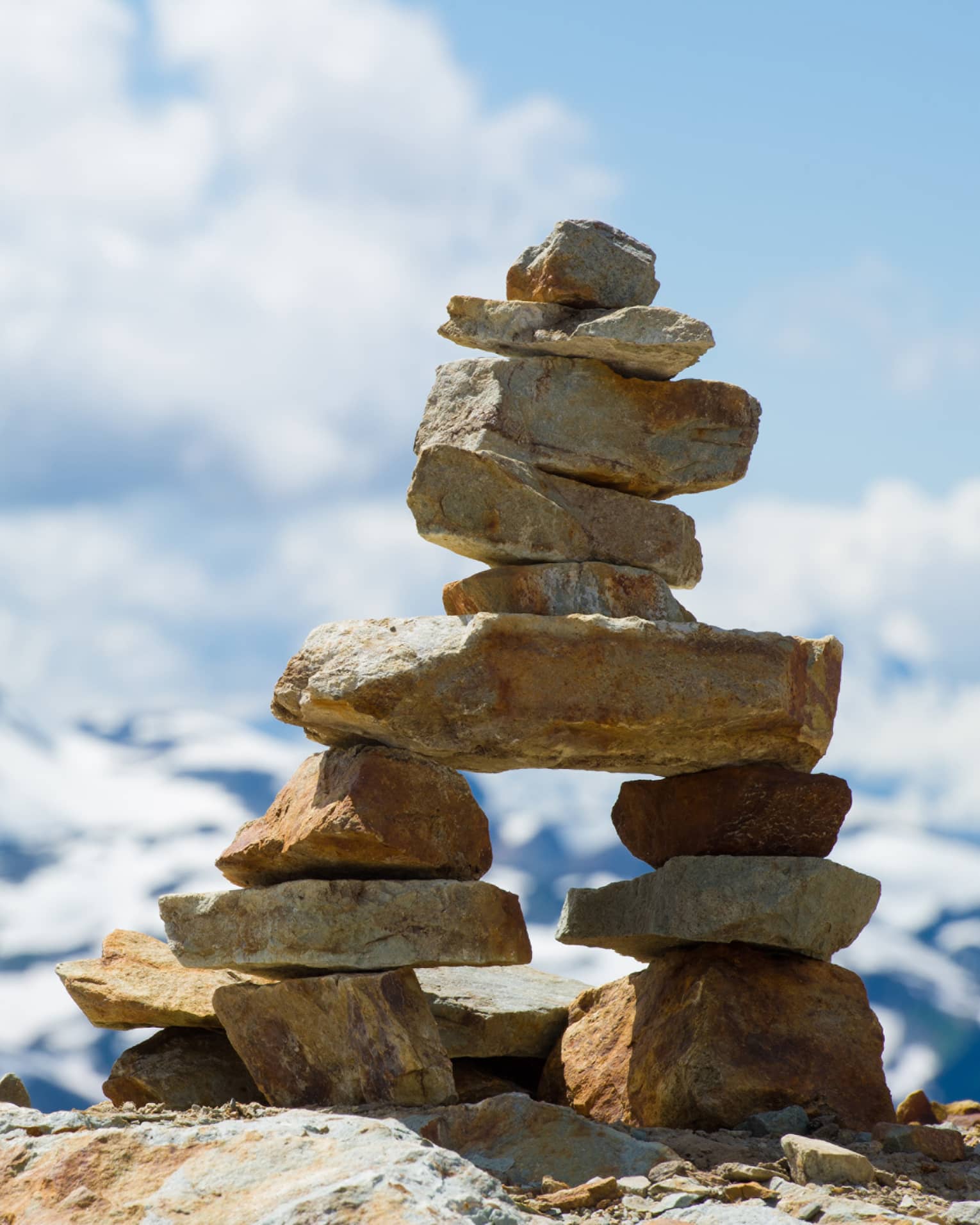 Close-up of an inukshuk atop a high rock shelf, against blue sky, white clouds and a majestic distant mountain.