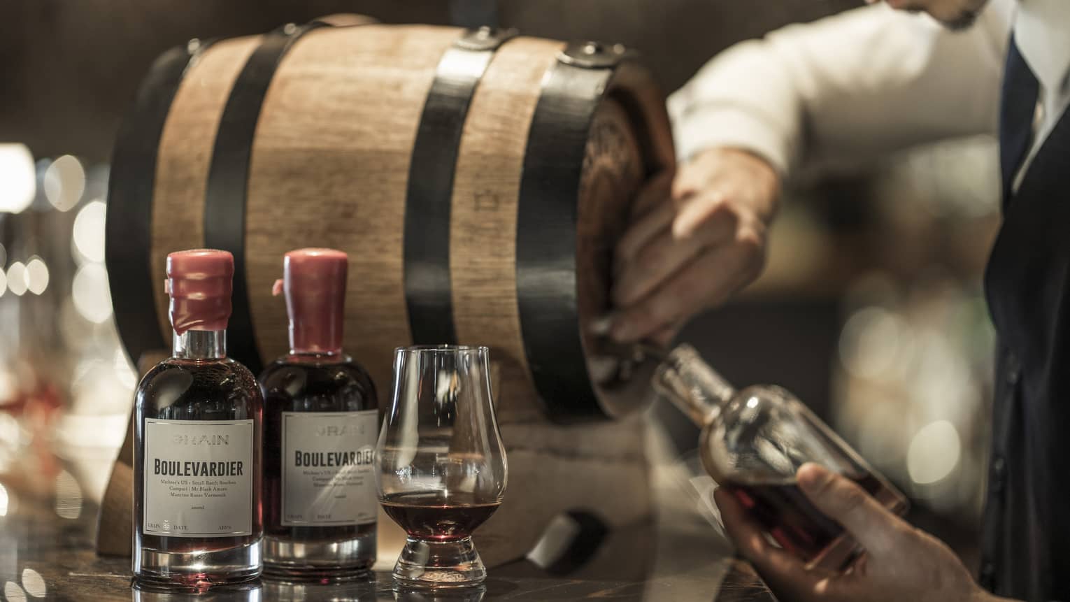 Bartender pours whisky from small barrel into bottle beside liquor bottles, glass