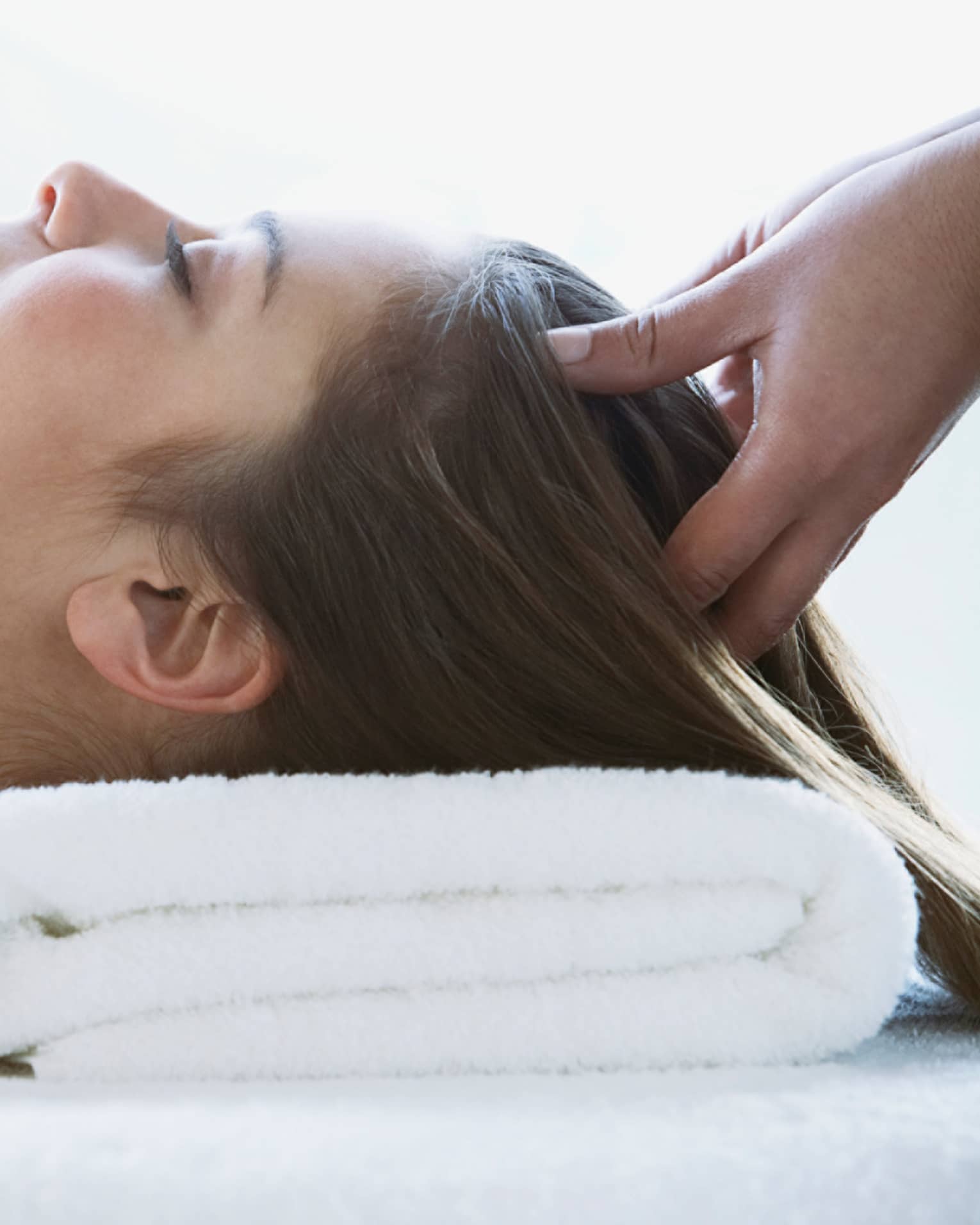 Woman lays on back with head on folded white towel, closes eyes as hands massage her scalp