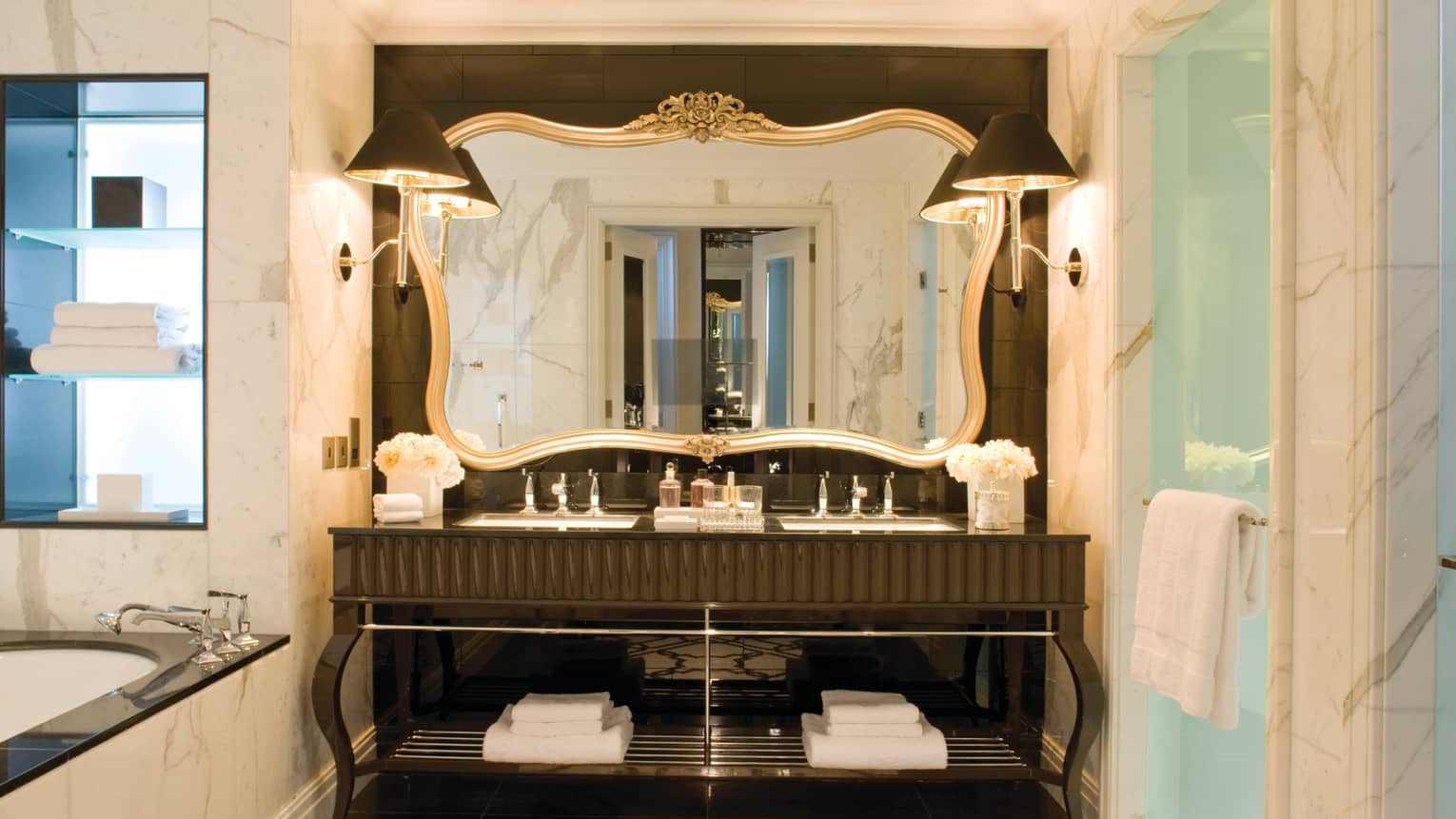 Large hotel bathroom vanity with double sink, tall mirror with gold frame, corner of marble bath visible