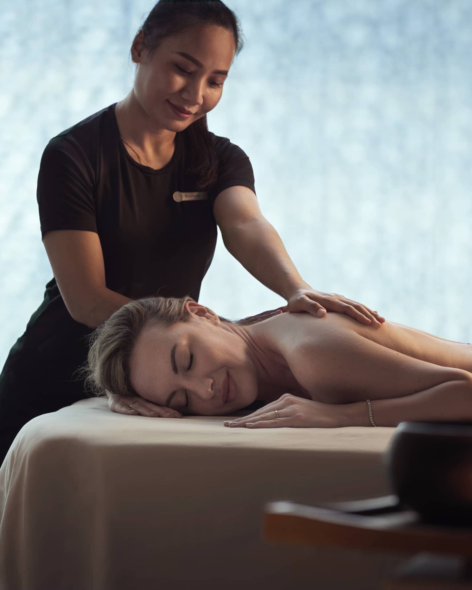 Spa staff massages woman's shoulders as she lays on bed in dark room
