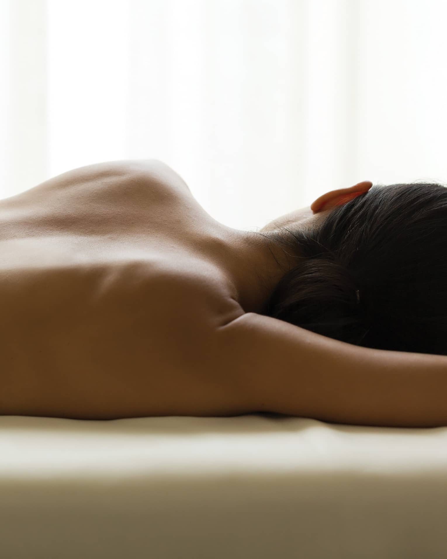 Woman's bare back as she lays on massage table under window, white curtains