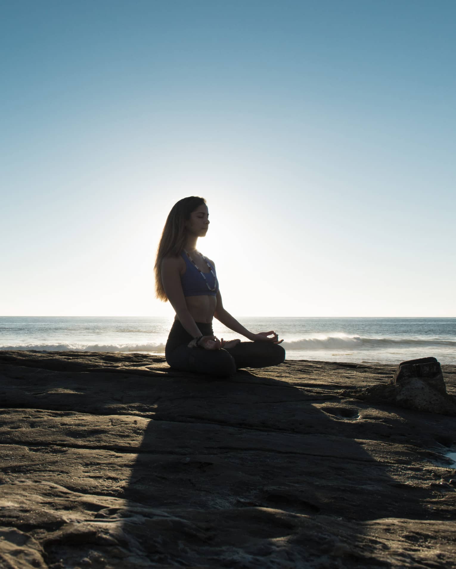 Silhouette of woman sitting cross-legged, meditating on rock by ocean