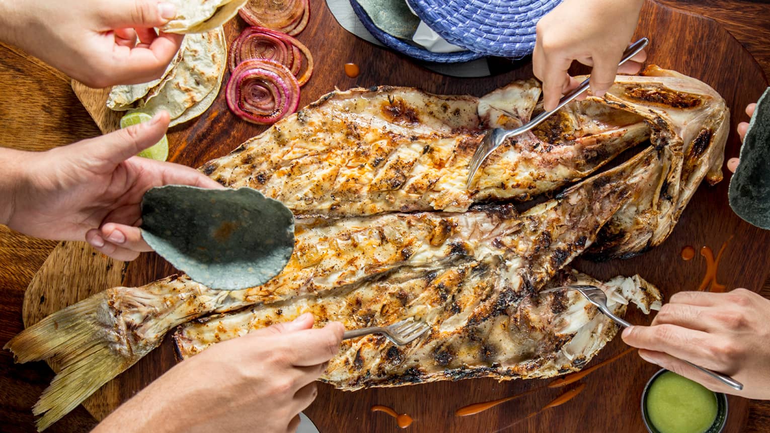 Aerial view of table, hands holding forks, digging into whole grilled fish on platter