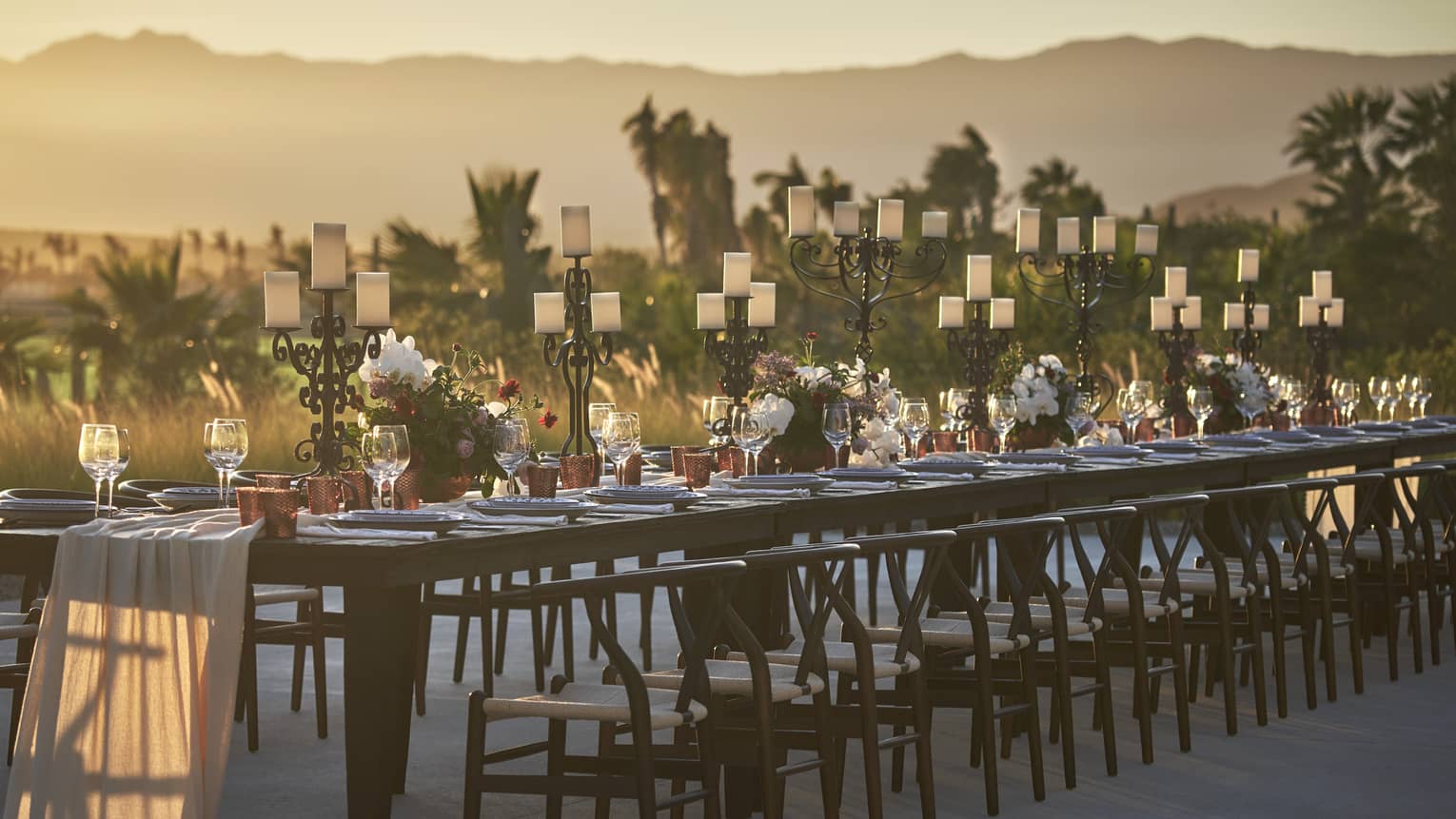 A long dark wood rectangular table is set with a white cloth draped down the center, white dishes and red accents lining the top, dark mid century modern dining chairs tucked in on either side while the sun sets over the mountains in the background