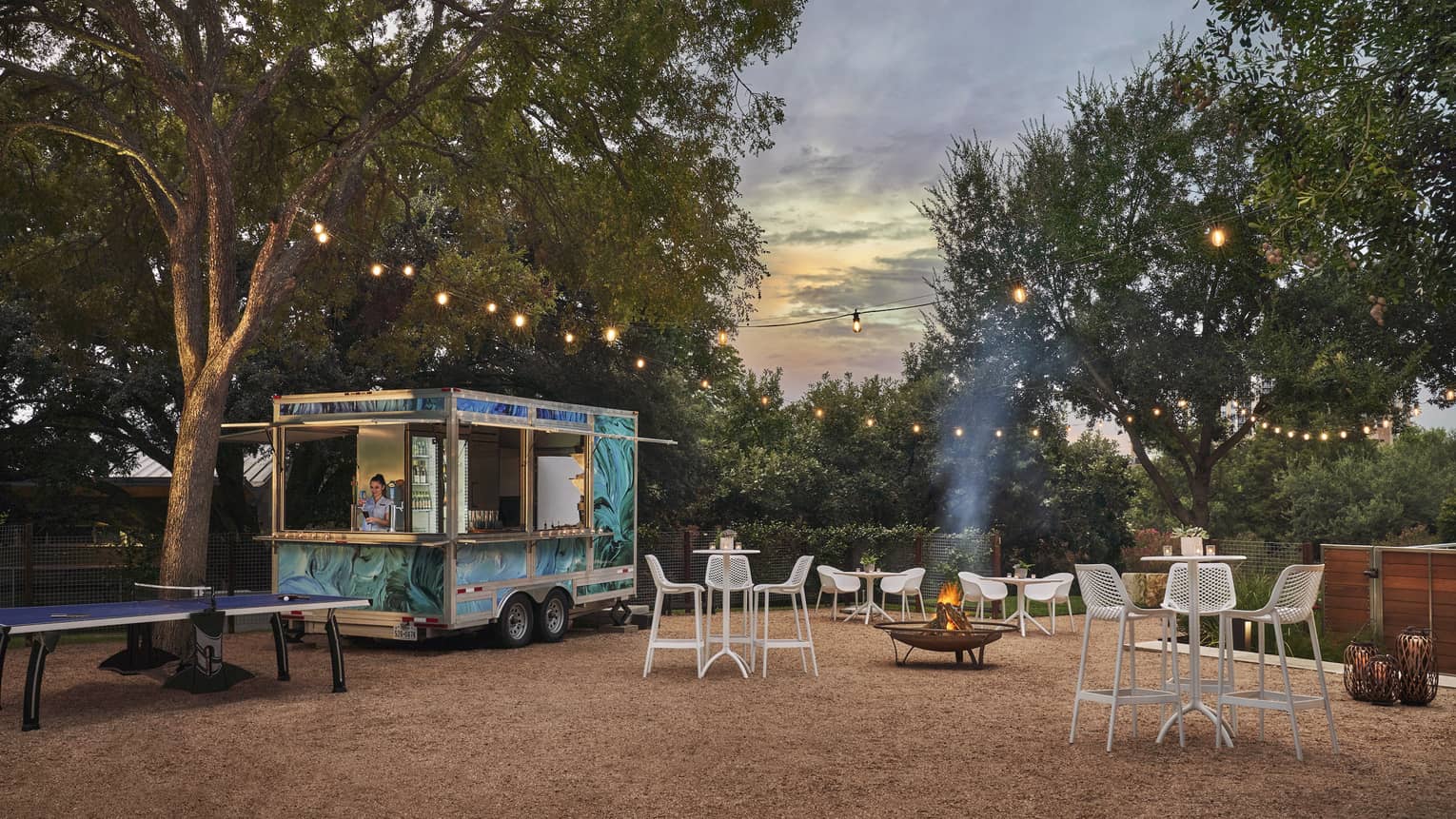 A food truck outsider surrounded by chairs, tables and trees.
