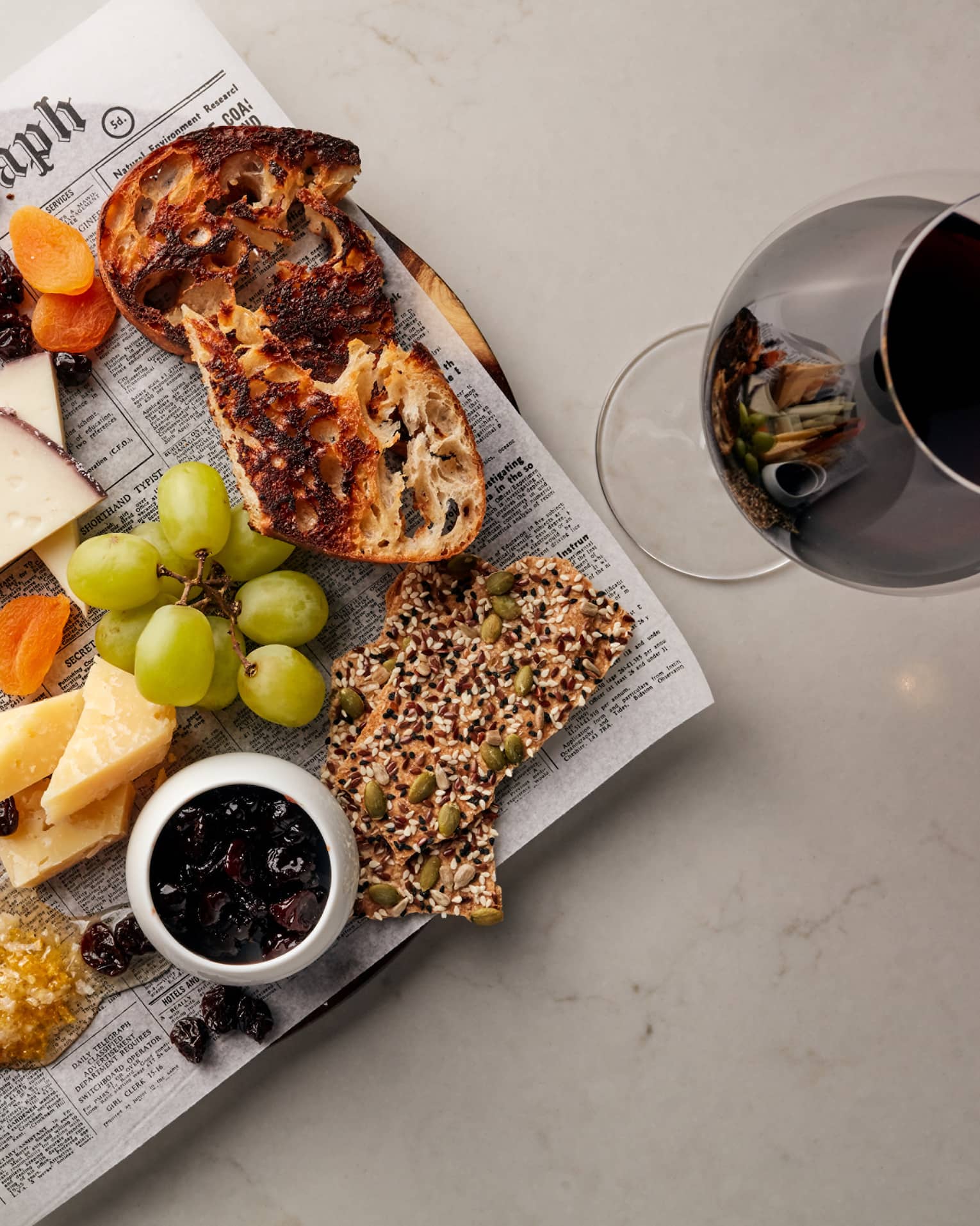 Cheese, grapes, crackers and bread on newspaper on platter next to glass of red wine