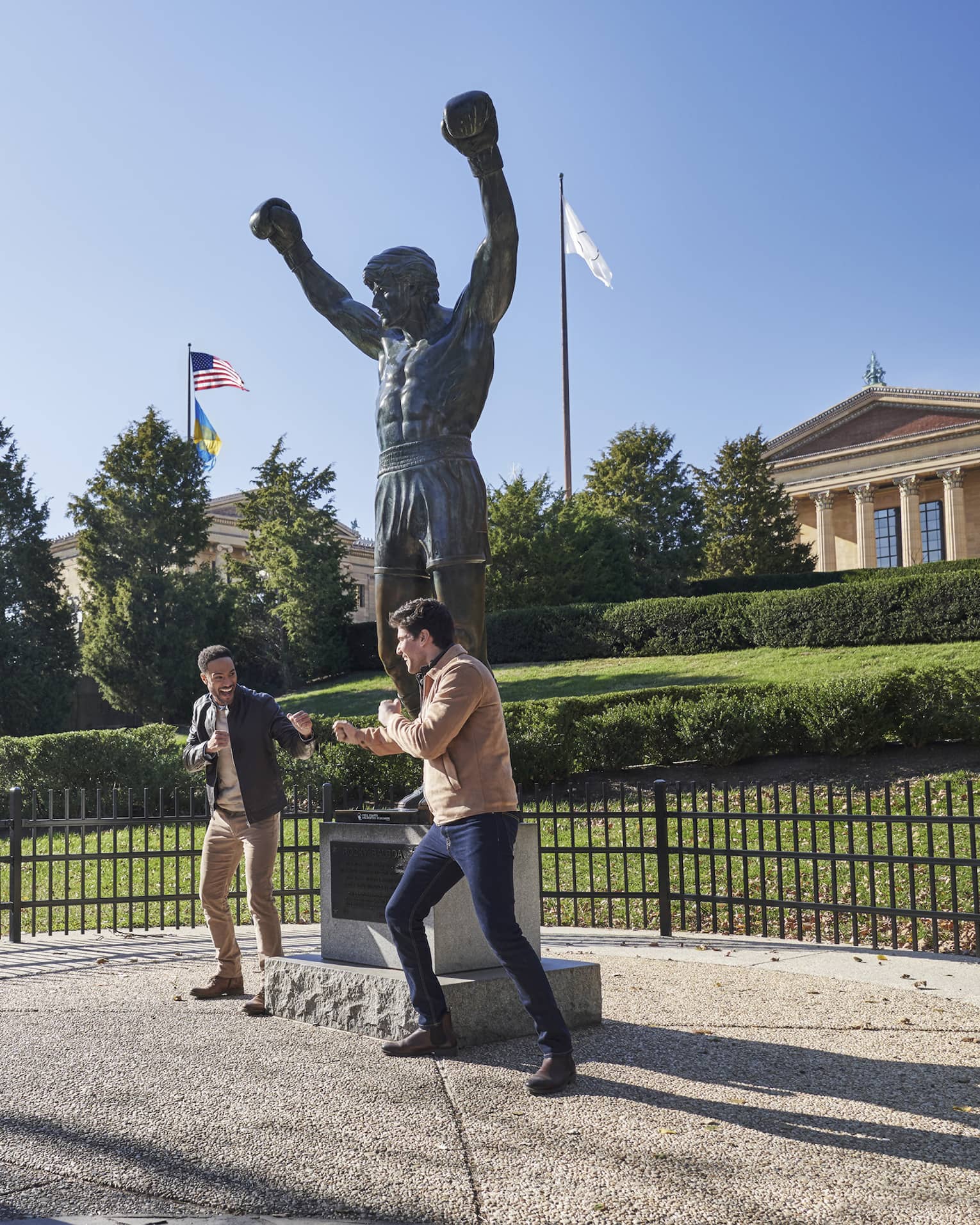 Two people pretend to box in front of the Rocky statue against a bright blue sky; another takes their photo, laughing.