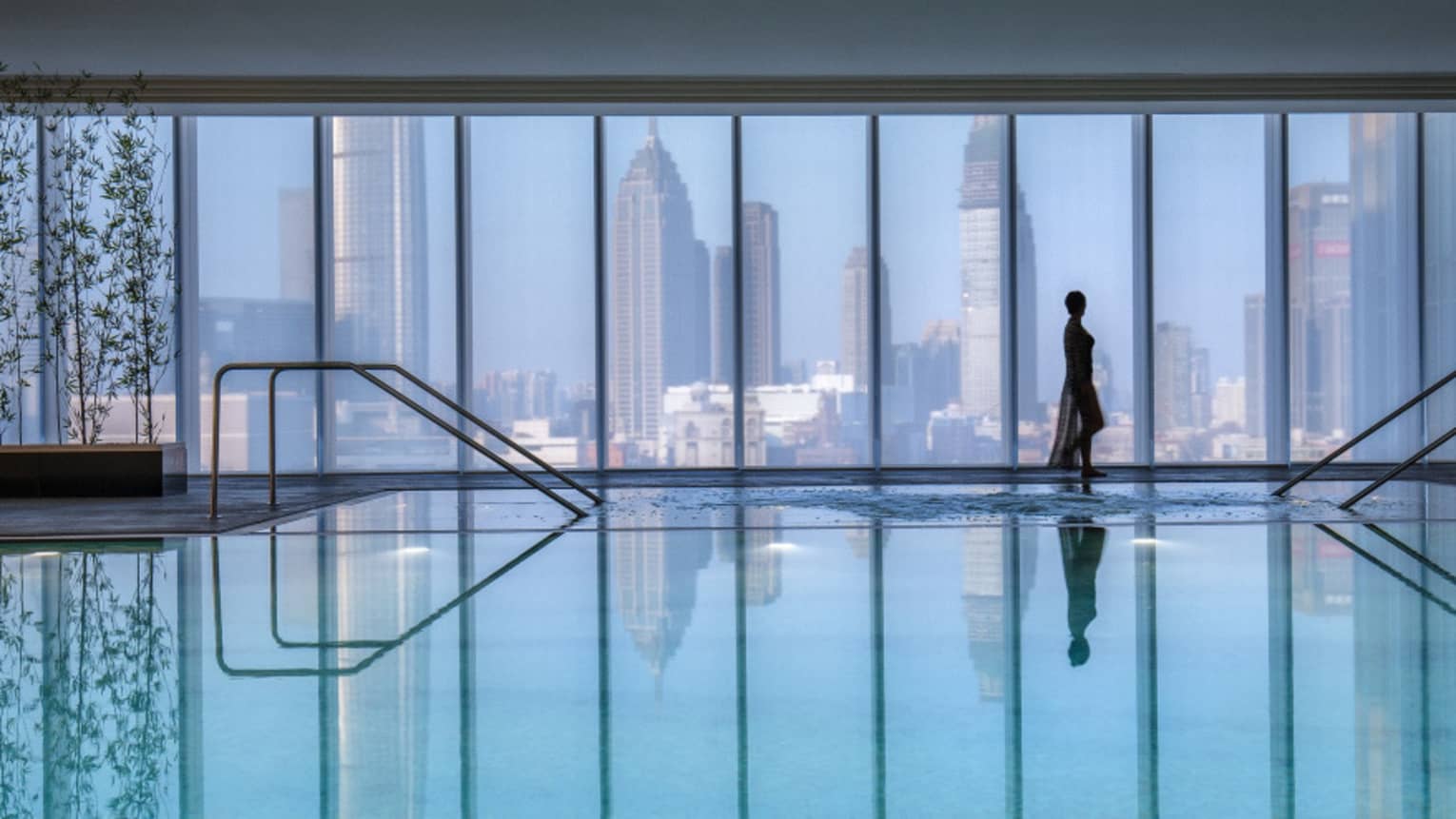 Silhouette of woman walking by blue indoor swimming pool, floor-to-ceiling window