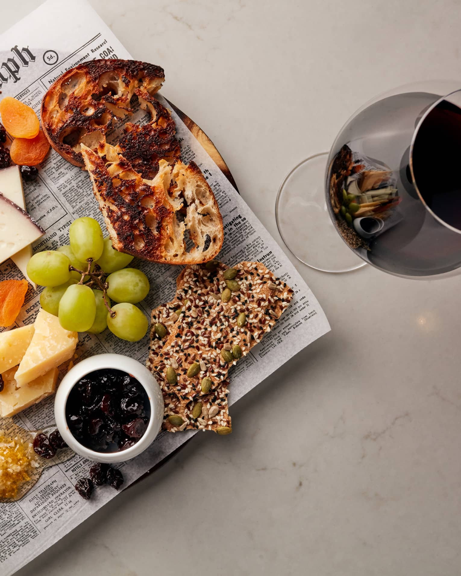 Cheese, grapes, crackers and bread on newspaper on platter next to glass of red wine