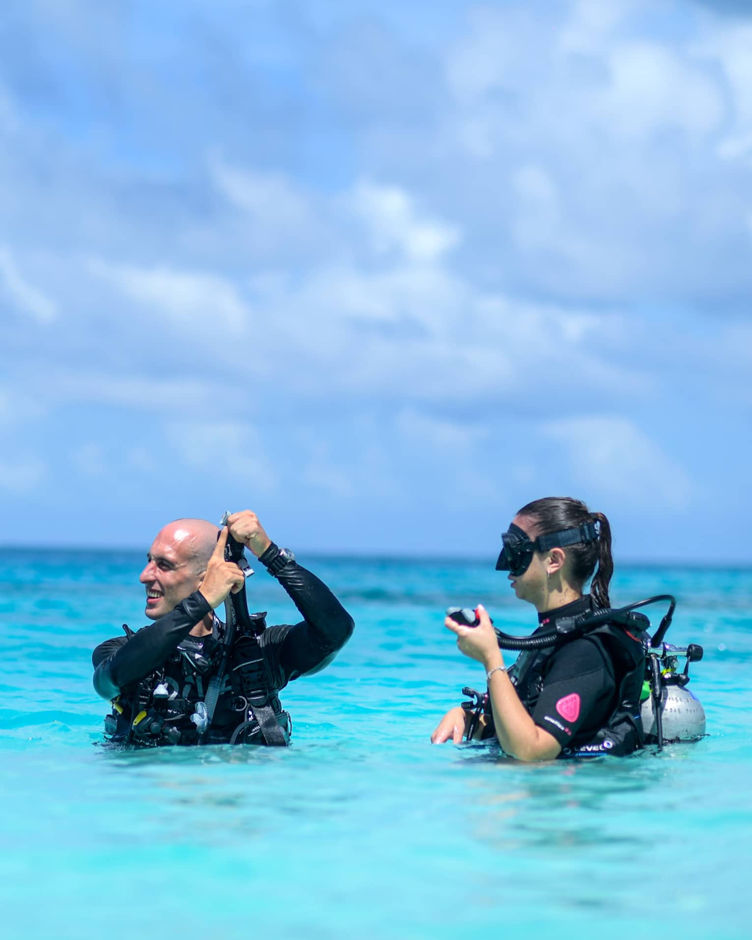 Scuba instructor showing mouthpiece to guest as they stand wearing scuba gear, chest-deep in azure water under fluffy clouds.