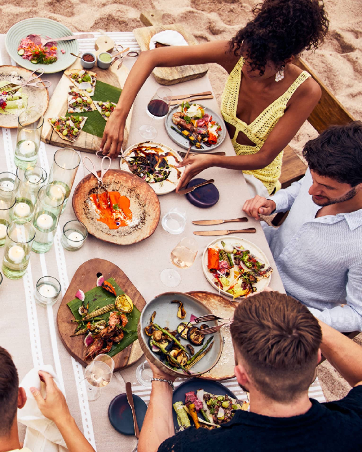 A group of people eating various food on a large table at the beach.