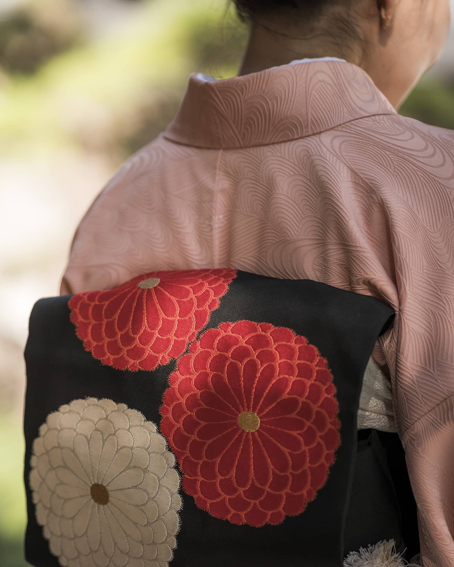 Close-up rear view of a person in a pink kimono with graphic pink and white chrysanthemums on the attached black obi cushion.