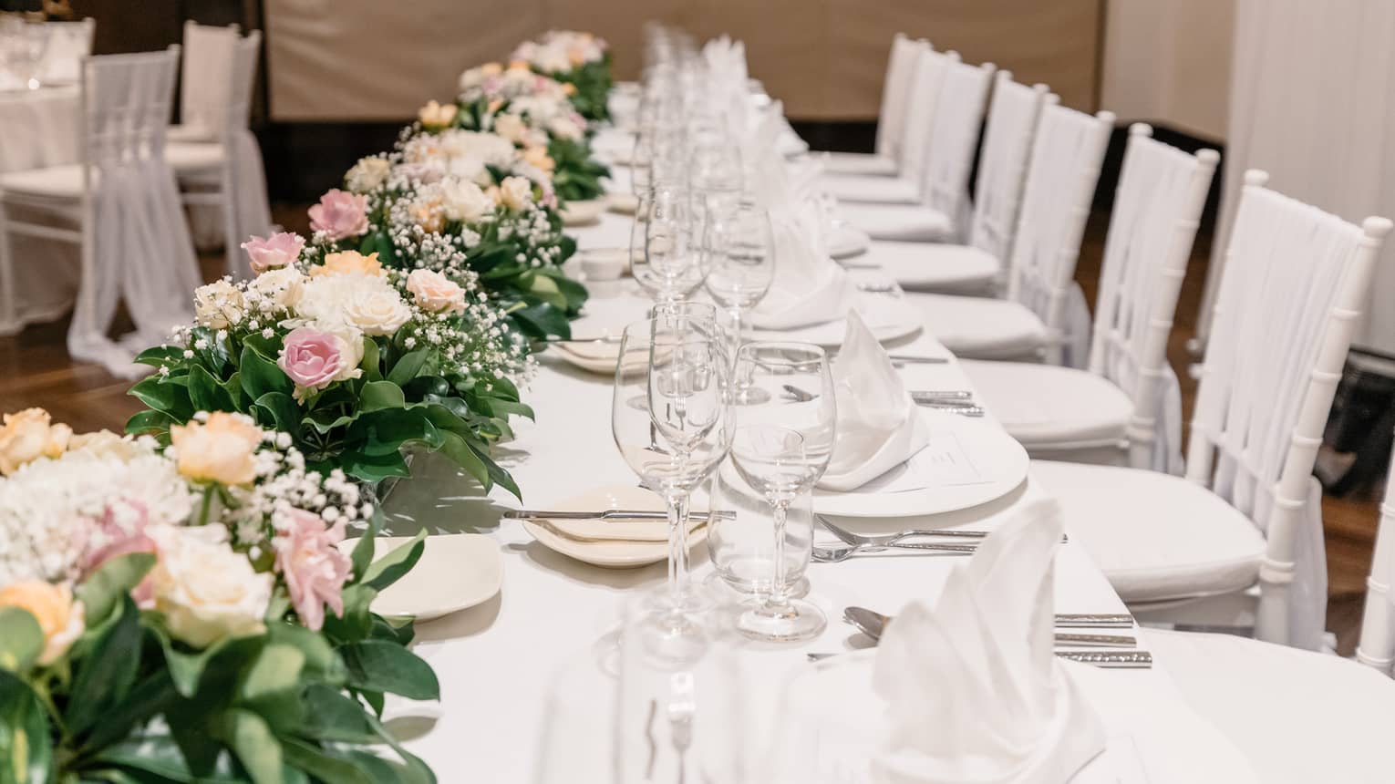 A banquet table is set with a white tablecloth, white porcelain dishes and clear glassware, green, white and pink florals