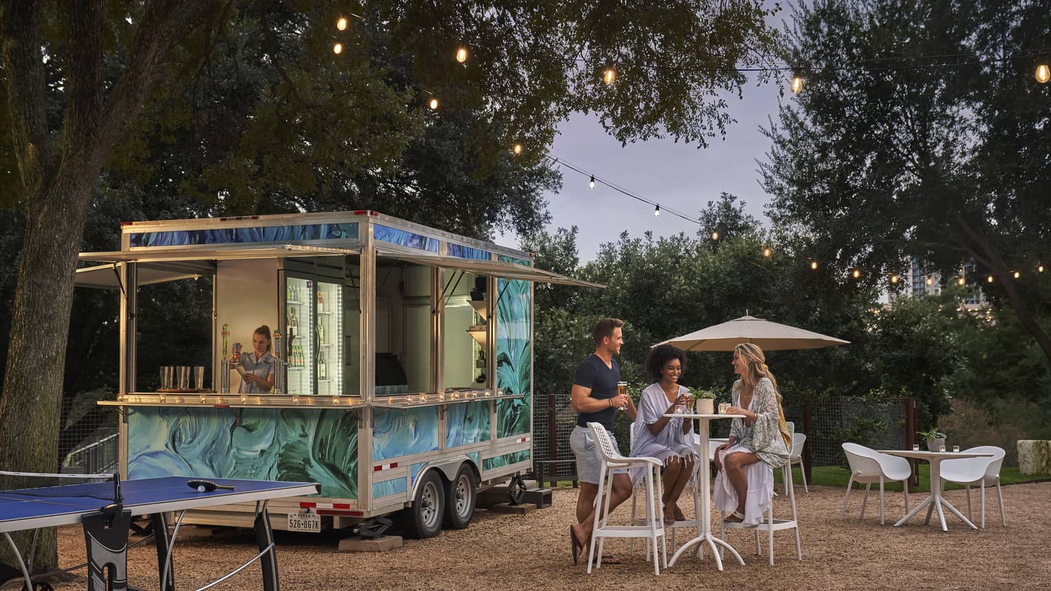 Three people sitting outside of a food truck surrounded by trees.