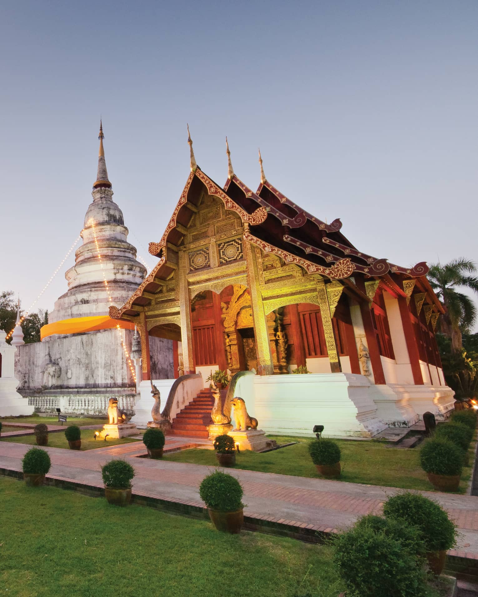 Two temples with gabled roofs and intricate carvings uplit in gold, between them a white domed building and stone monument.