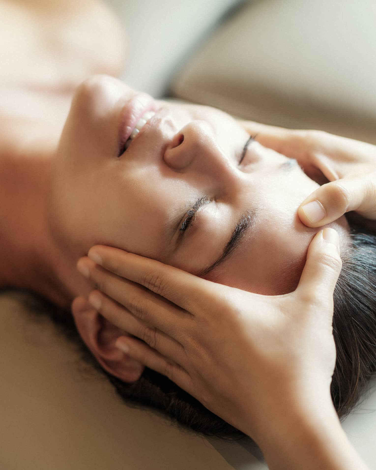 Spa facial, two hands rest on woman's forehead as she closes her eyes, lays under sheet on treatment table