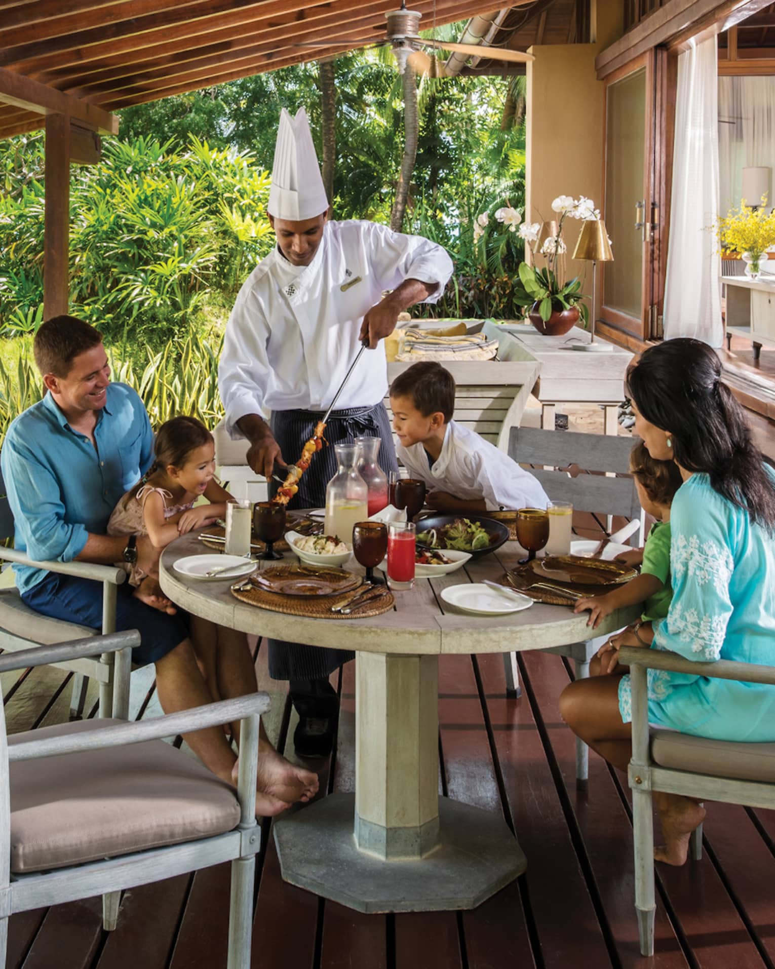 Chef in uniform presents meat skewer to family around table on Beach Villa patio