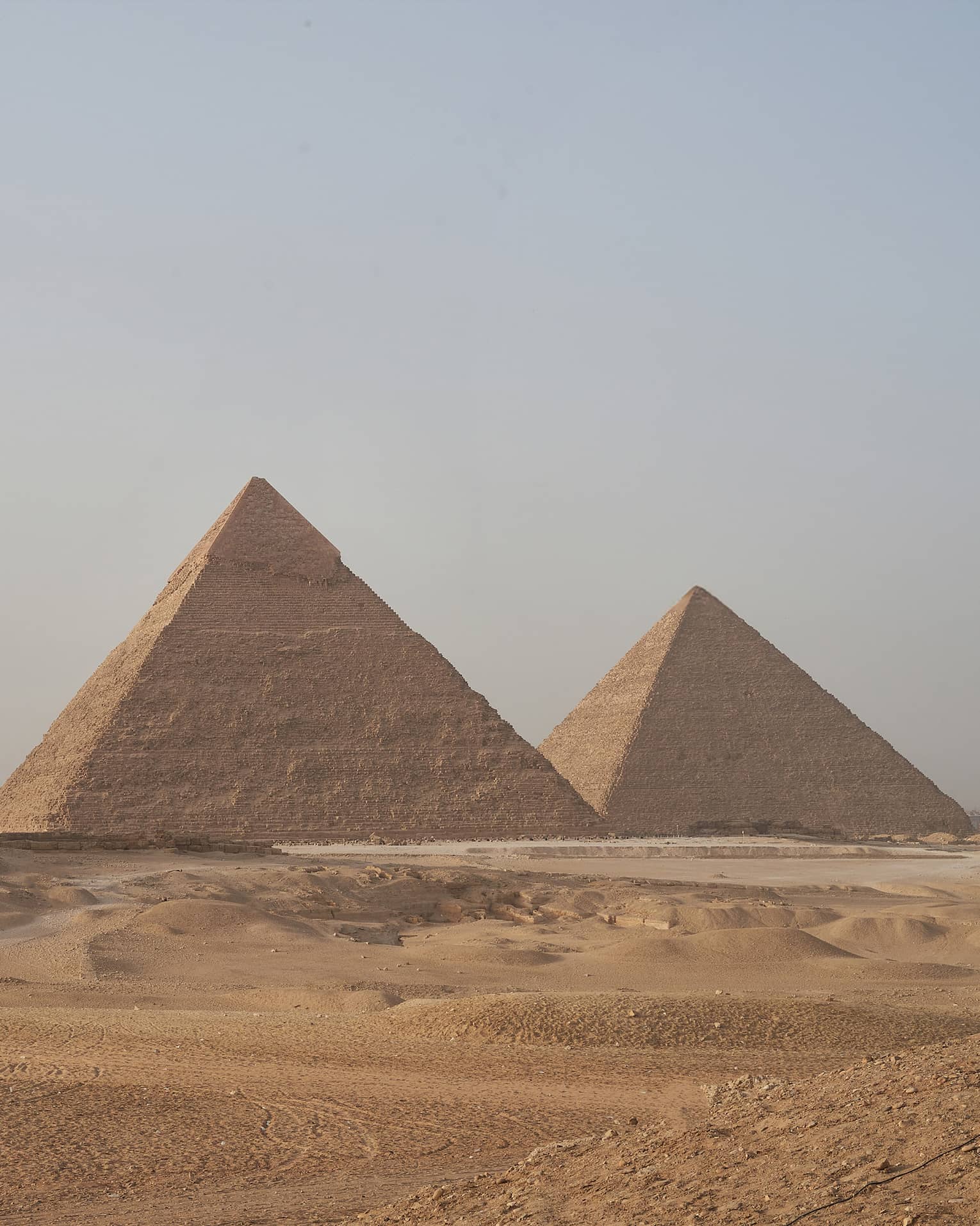 In the arid desert, a line of three towering pyramids dwarf two smaller pyramids before them, under a hazy blue sky. 