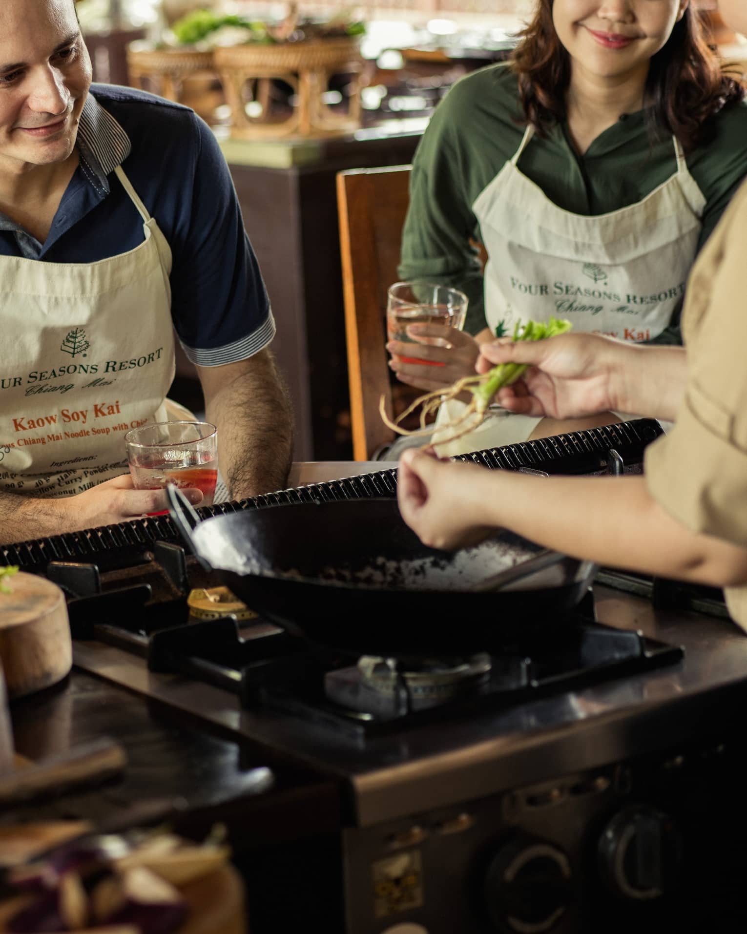 Three guests in aprons sit on stool in front of large wok, chef during Thai cooking class