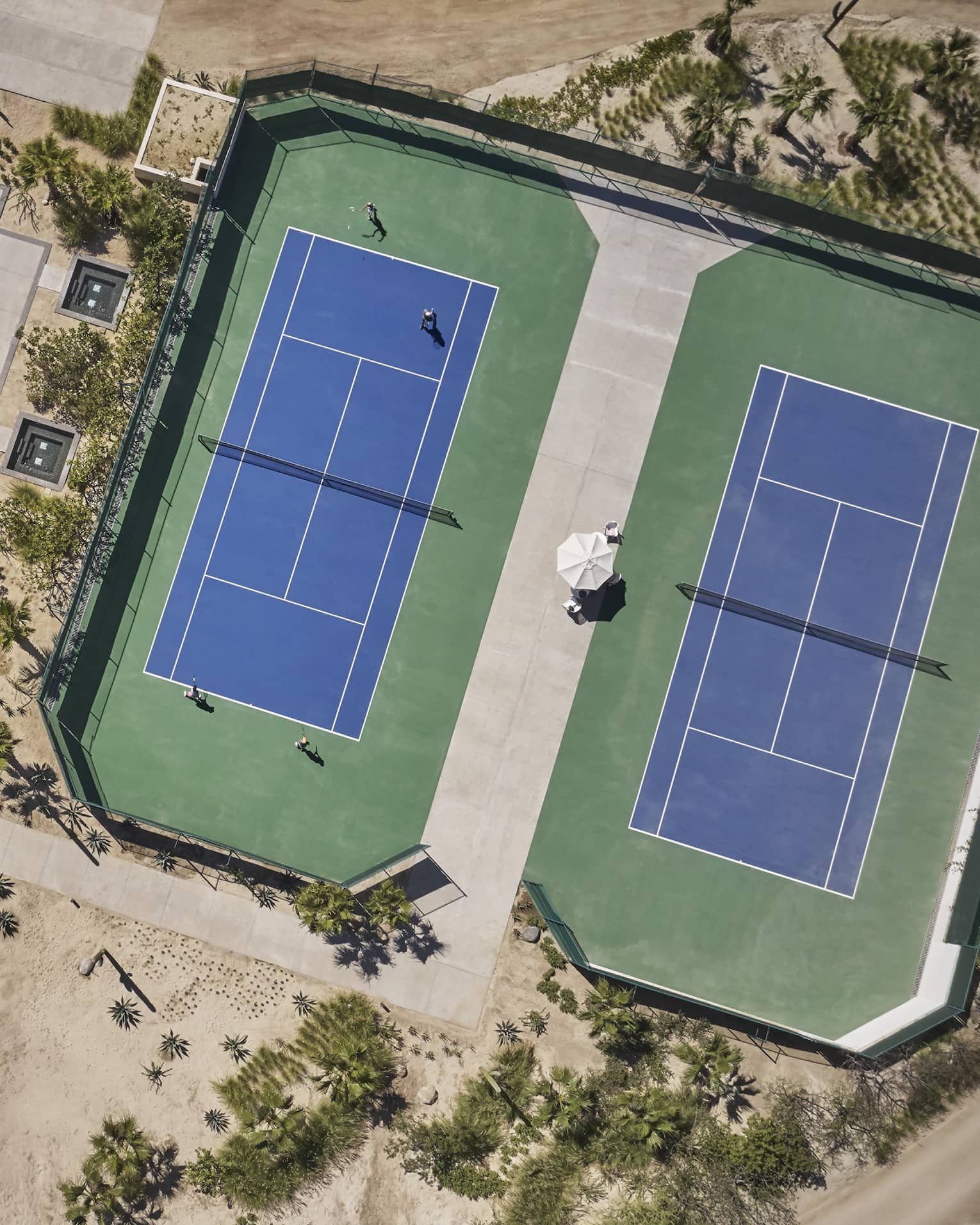 Aerial view of two green and blue tennis courts, one long rectangular pool and one blue basketball court surrounded by sand and greenery