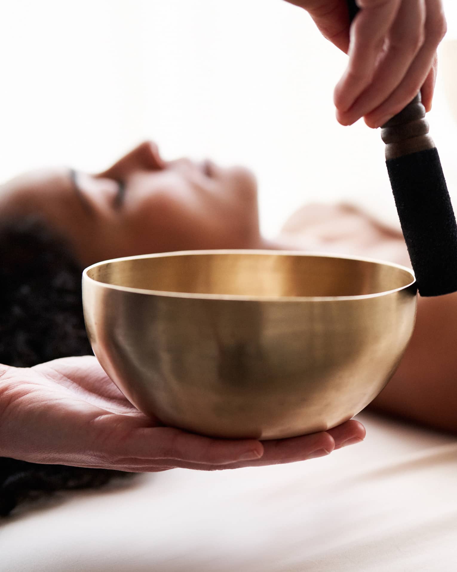 Close up of hands playing a singing bowl with a woman laying down on a spa treatment table in the background