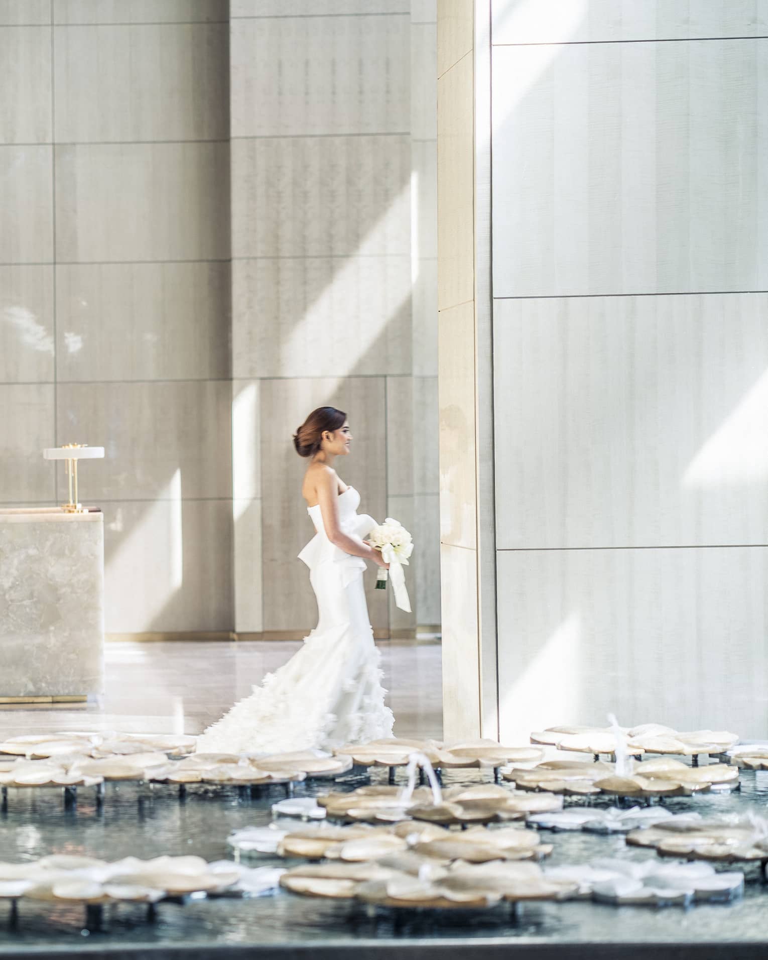Sun rays illuminates a bride as she walks through a white walled lobby carrying her bouquet of white flowers