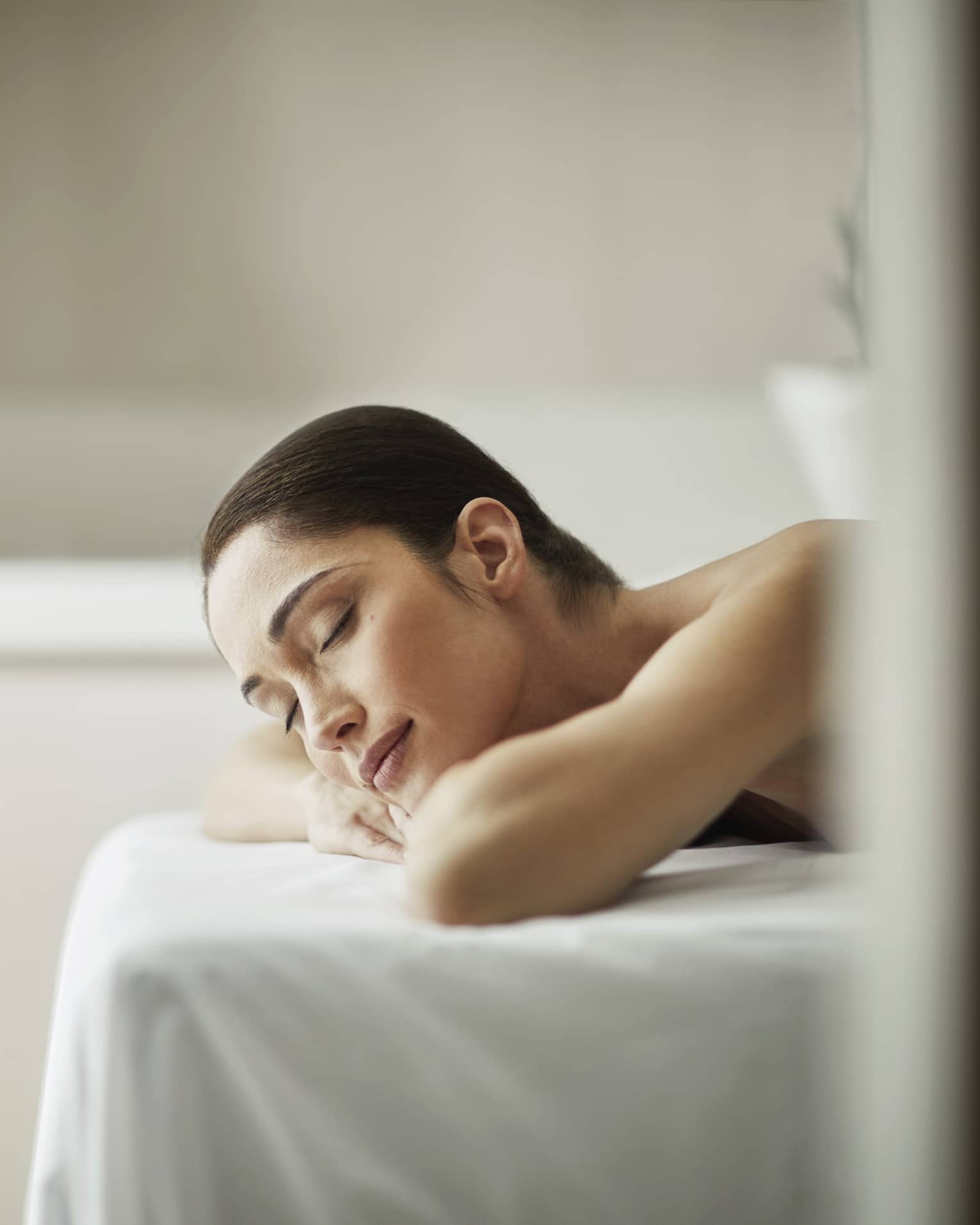 Woman smiles with eyes closed, rests head on arms as she lies on massage table with white sheet