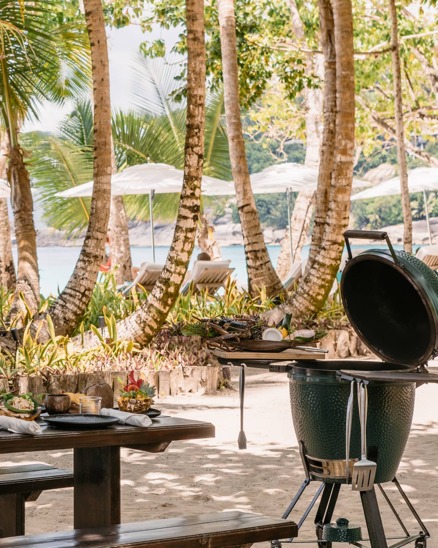 BBQ and grill equipment outside near a table and palm trees.