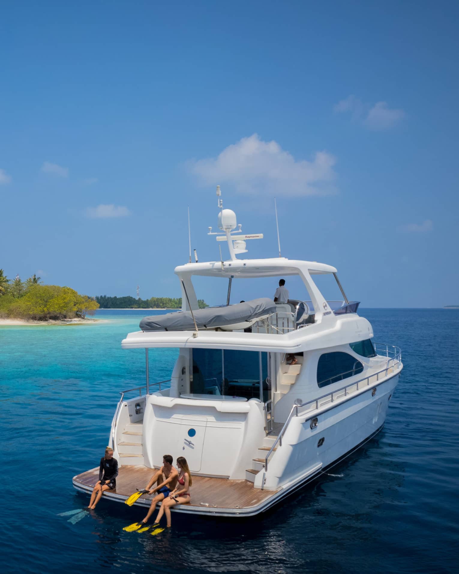 A couple sit with a marine biologist at the rear of a yacht, preparing to snorkel in smooth ocean water amid lush islands.