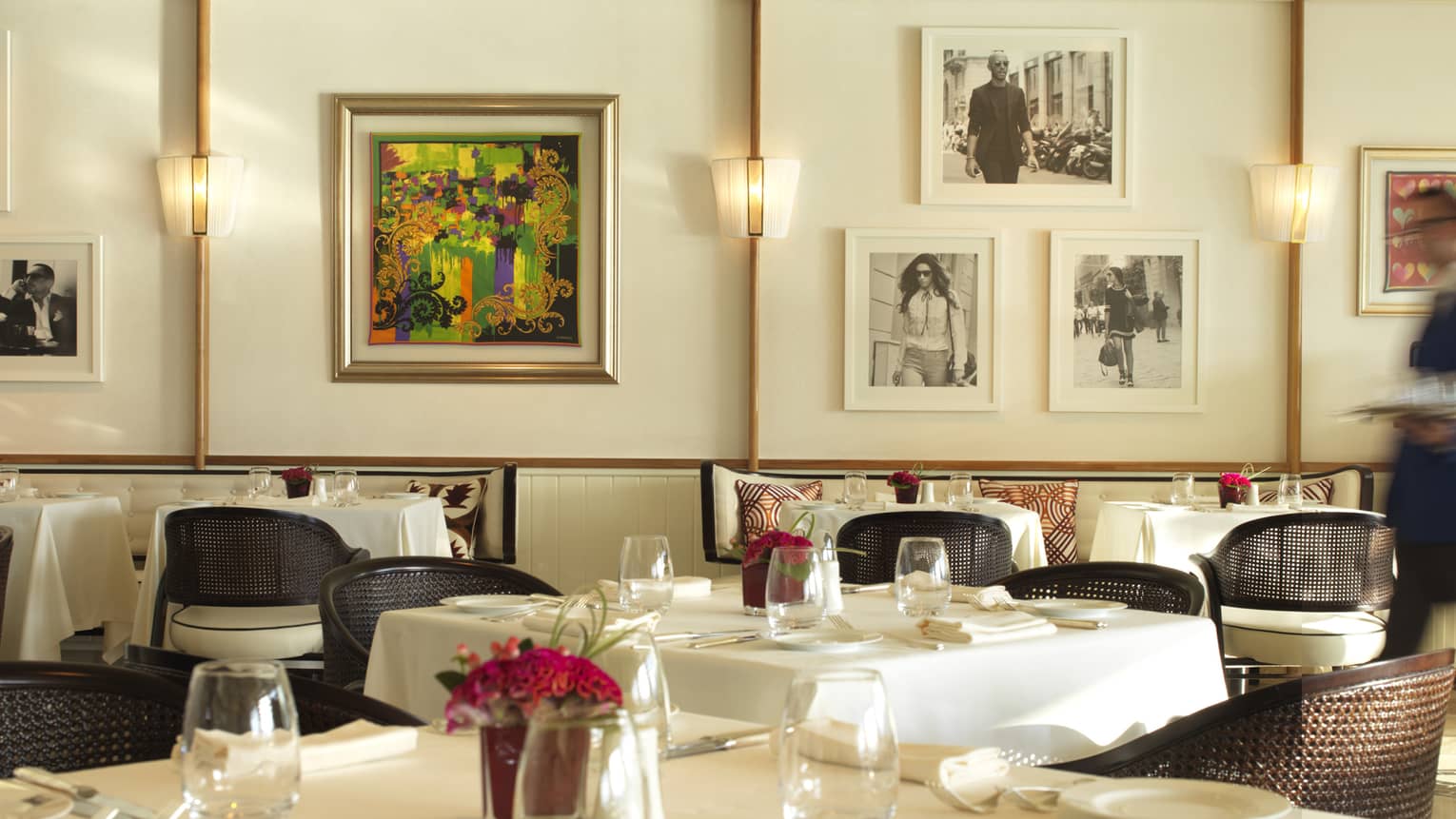 Server in blue suit walks through Cafe Milano dining room with elegant white dining tables, colourful art, framed black-and-white photos
