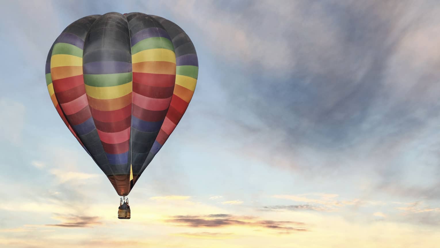 Rainbow-striped hot air balloon floats in the sky at sunset