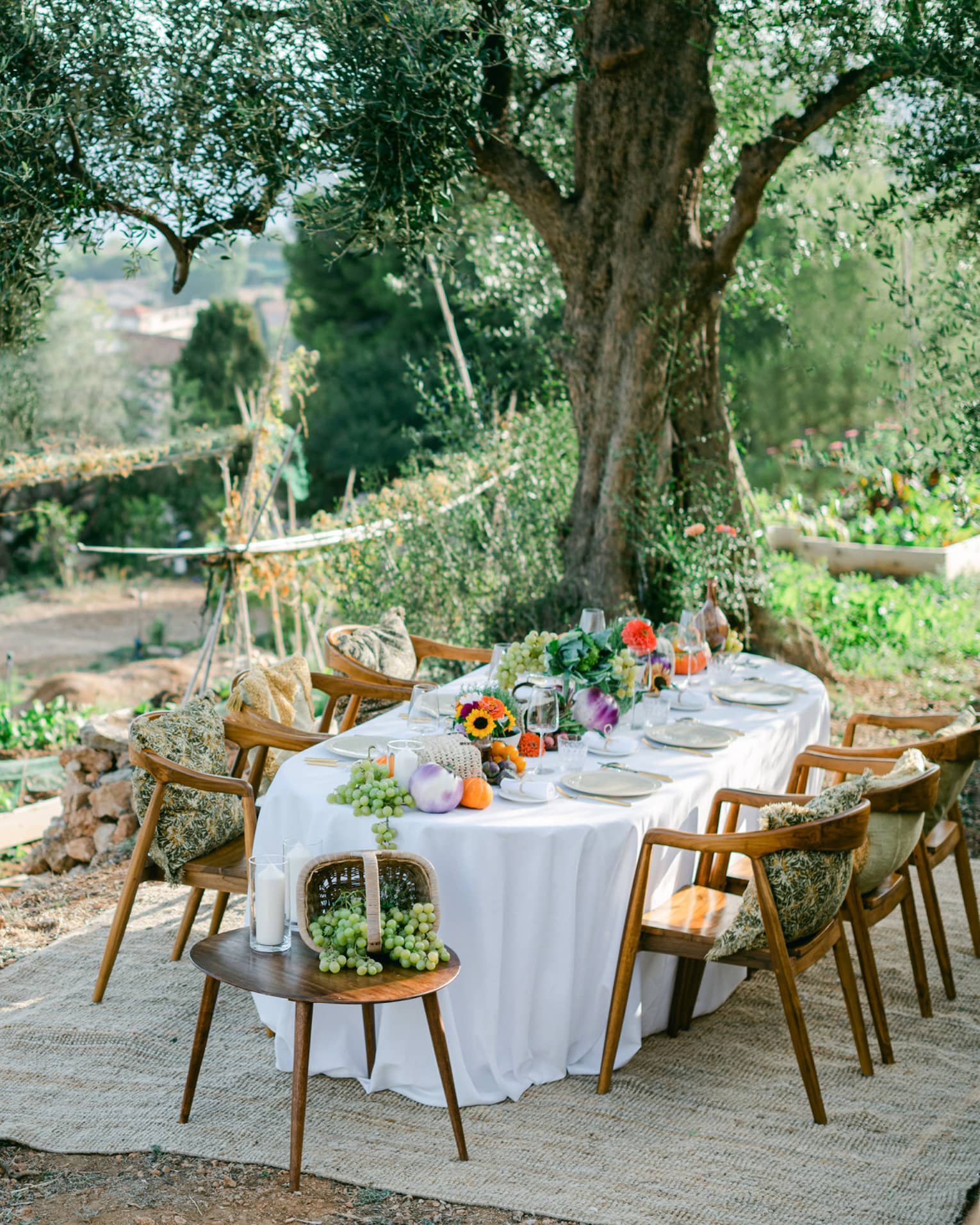 Garden dining setup with oval table draped in white and garden bounty down centre, and six chairs with pillows