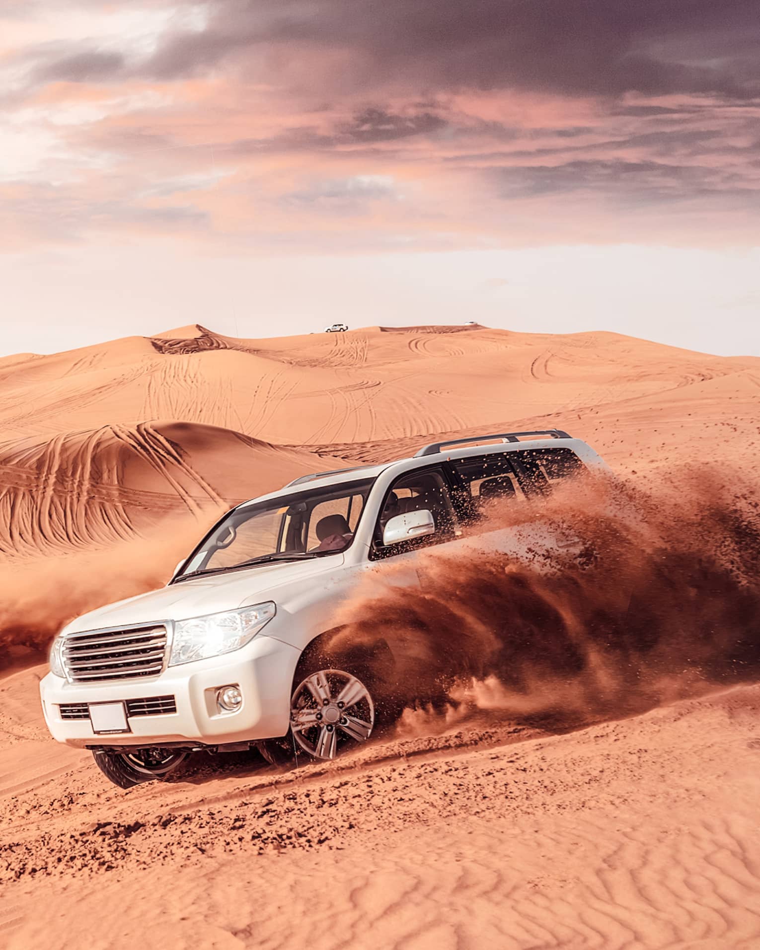 Sand flies from tires of two white SUV's driving across dunes