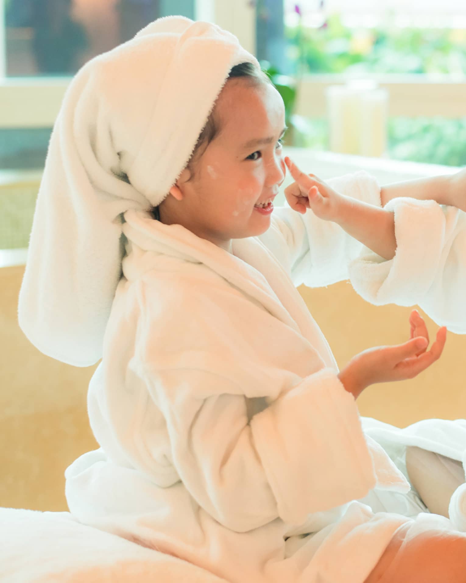 Two laughing young children in white bathrobes with towels around hair put lotion on each other's faces 