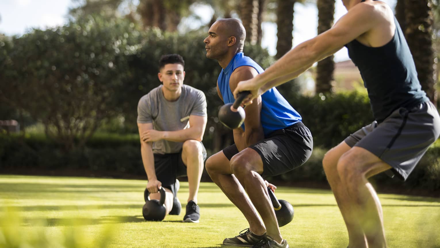 Four Seasons trainer watches as two men squat, lift kettlebell weights on lawn