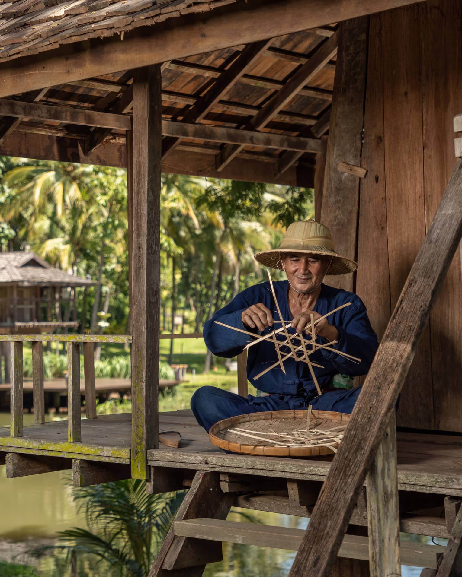 Man weaves a traditional Thai basket on the porch of a riverside pagoda