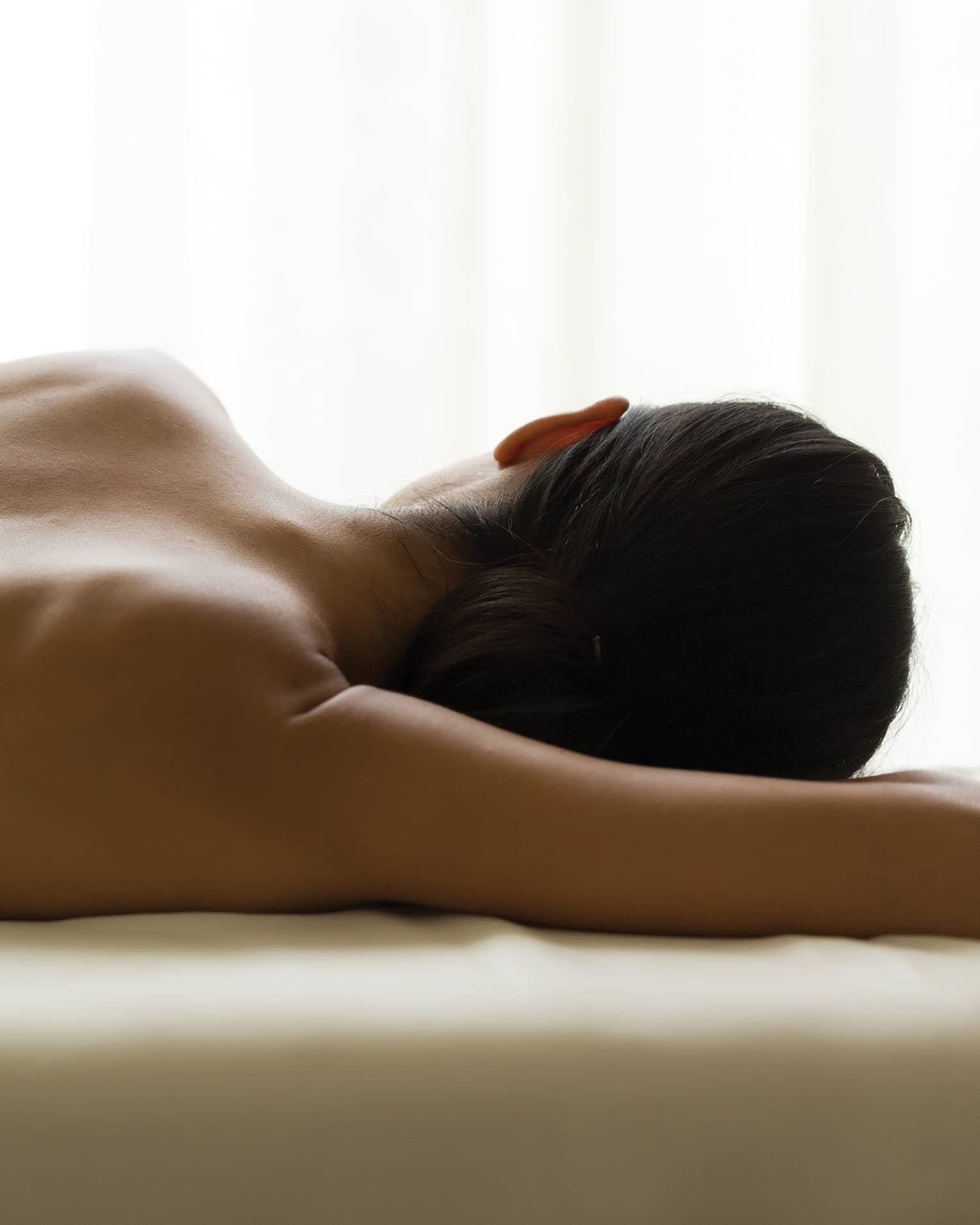 Woman's bare back as she lays on massage table under window, white curtains