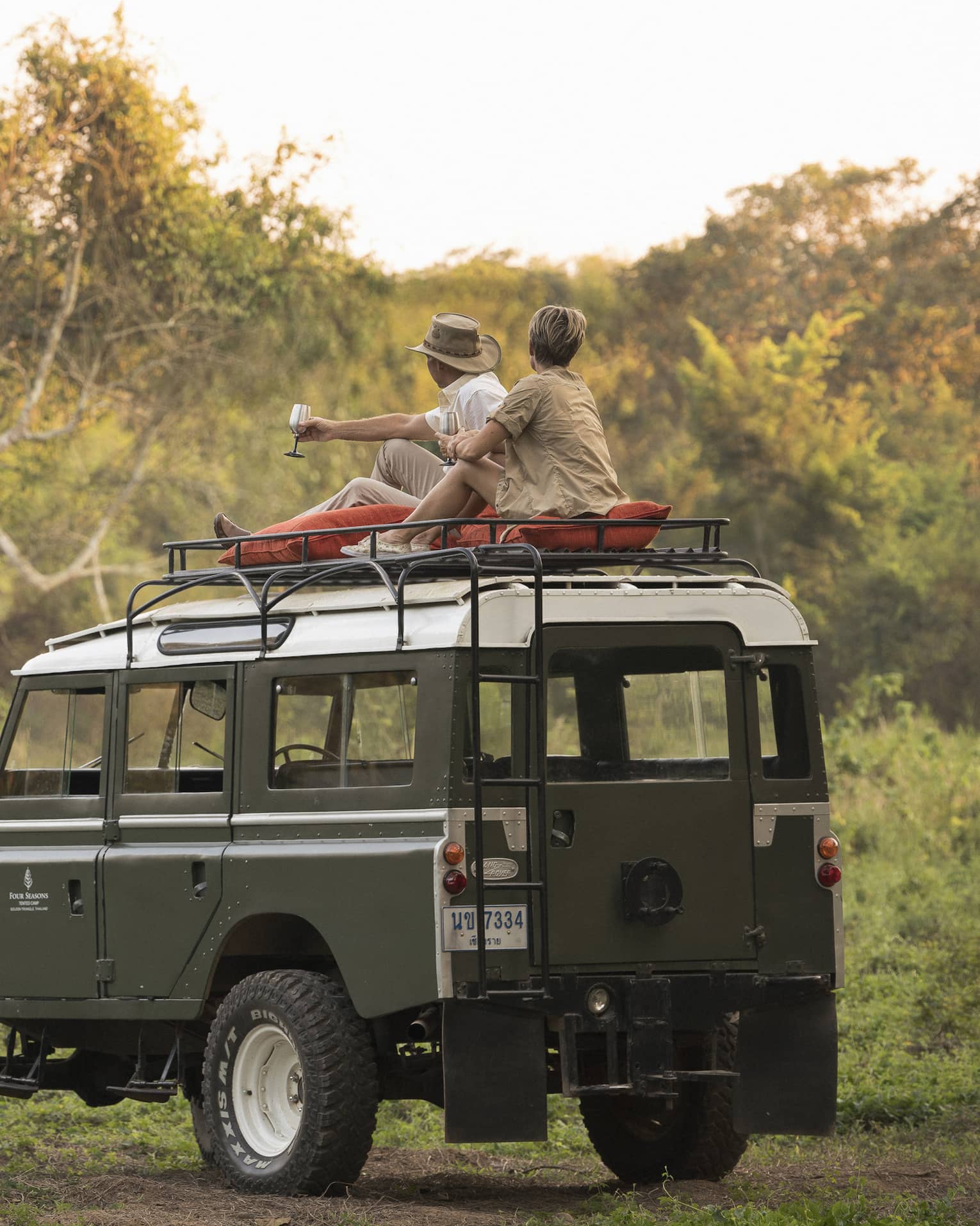 A couple sits on the roof of a Land Rover in the Bush, drinking a cocktail