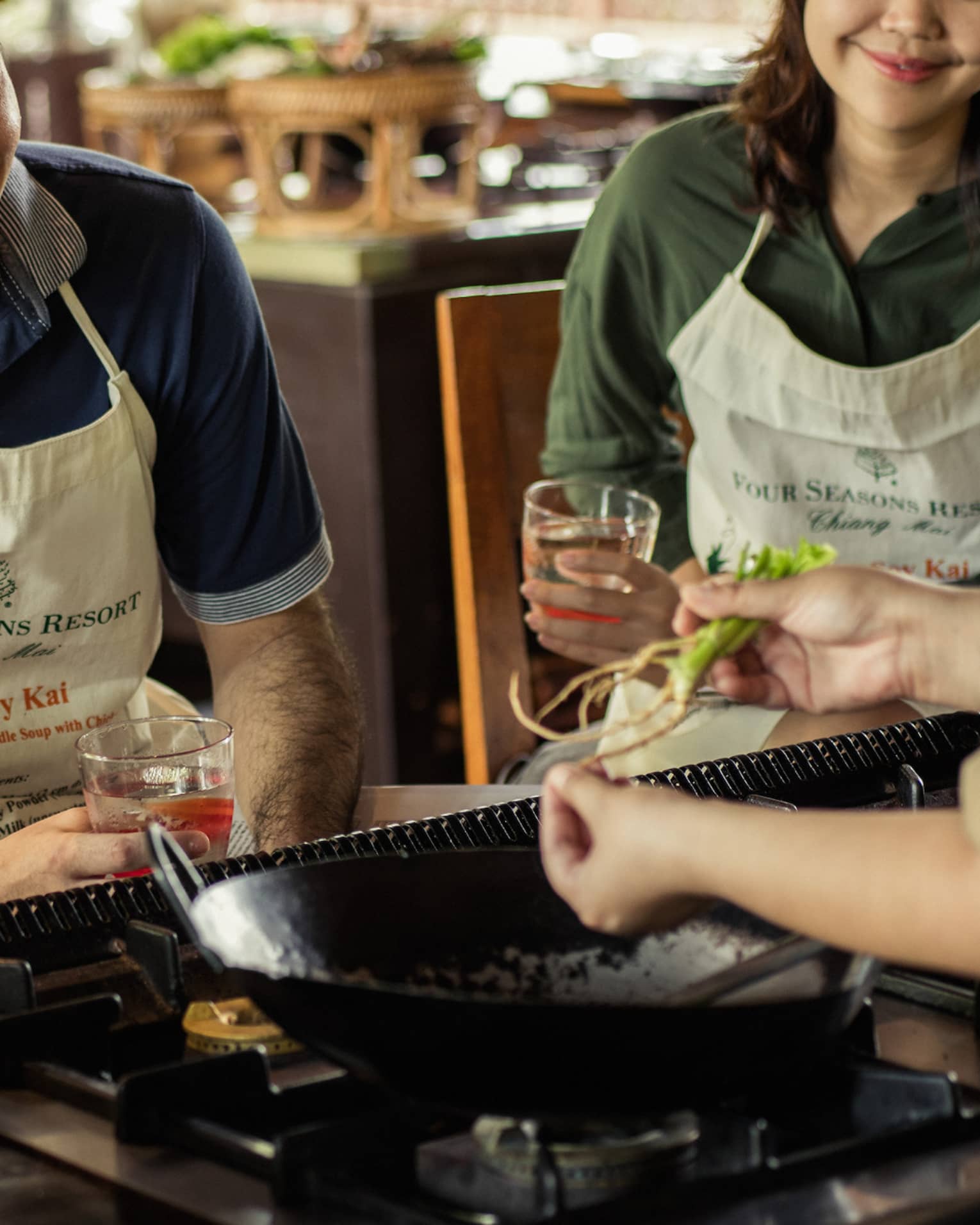 Three guests in aprons sit on stool in front of large wok, chef during Thai cooking class