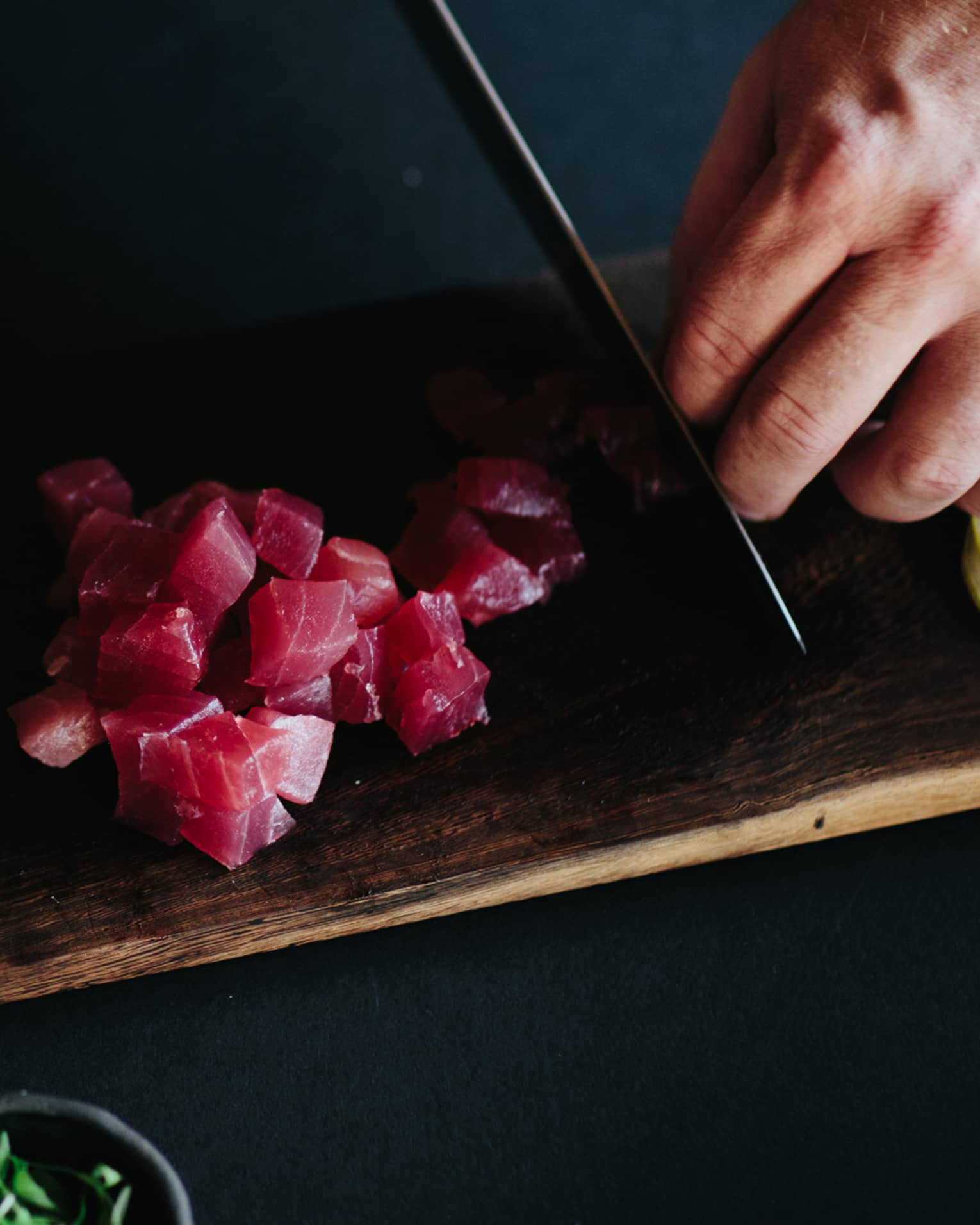 Man slicing raw tuna into cubes with knife on wood cutting board next to bowl of orange rock salt 