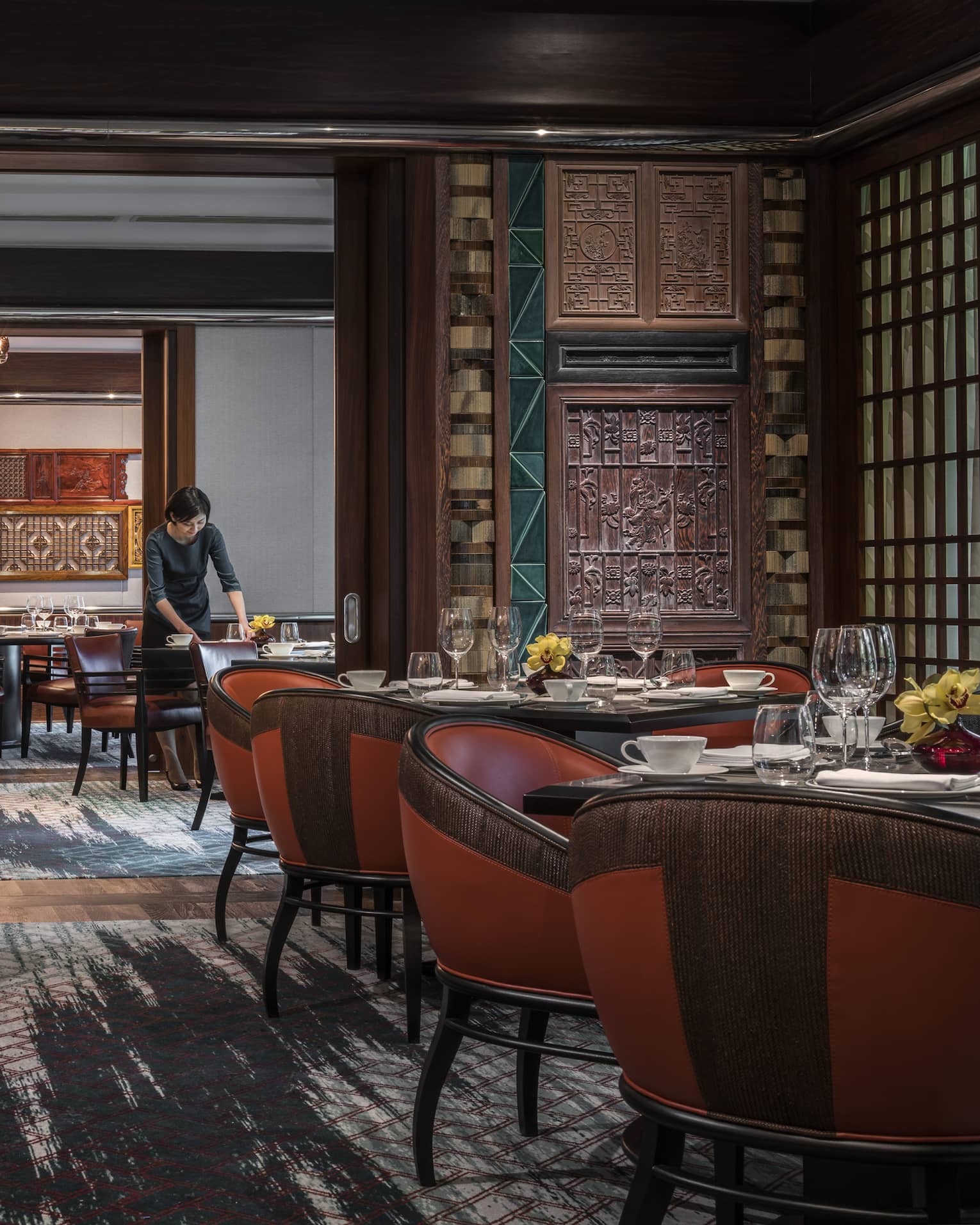 Woman sets table at Jiang-Nan Chun, Michelin-starred Cantonese restaurant, with red leather bucket chairs around dining tables, carved wood walls.